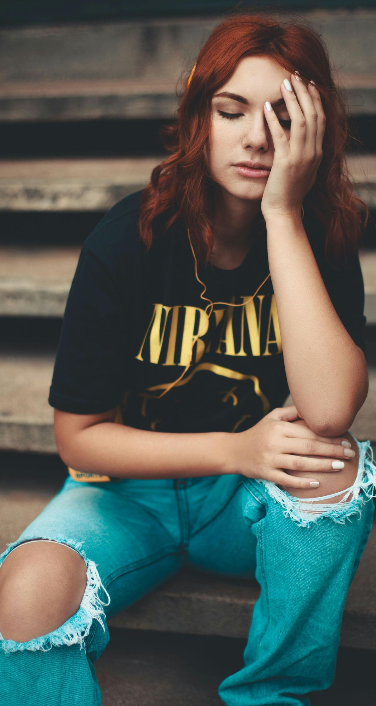 Person with red hair sits on steps, resting head in hand, wearing a black shirt and ripped blue jeans.
