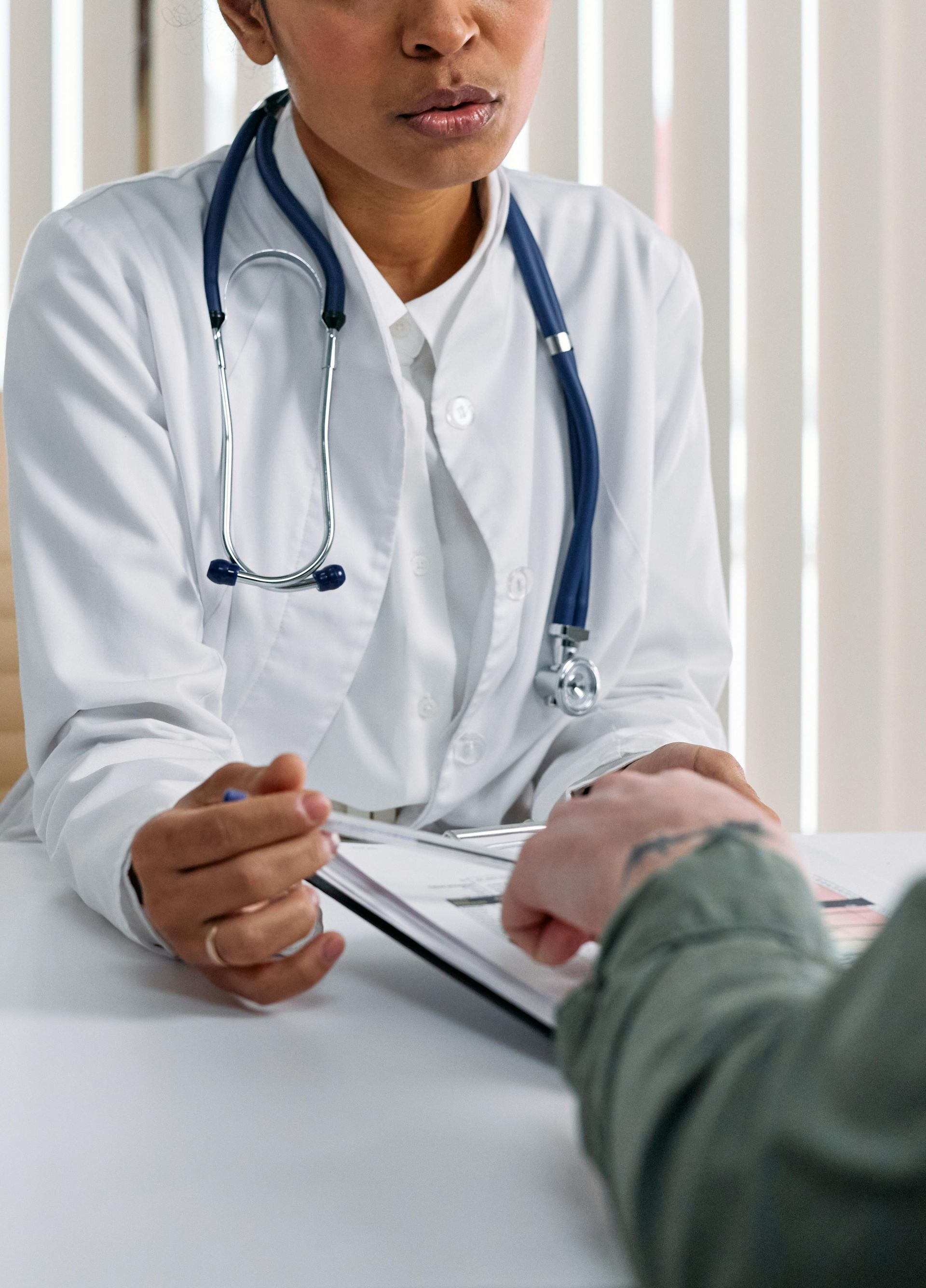 Doctor speaking with a patient across a desk, holding a clipboard and wearing a stethoscope.