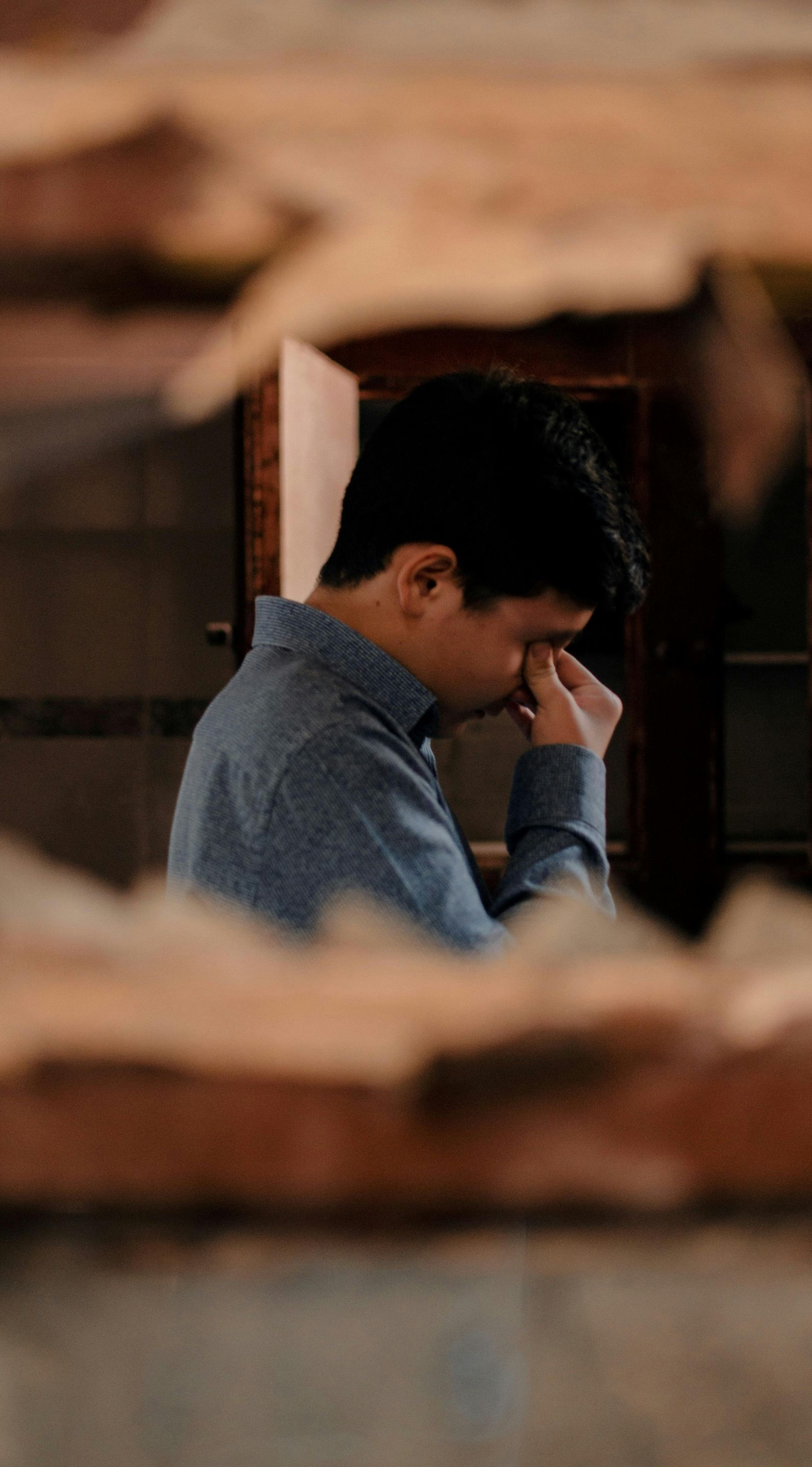 Person in a blue shirt sits indoors, resting their face in one hand, framed by blurred foreground.