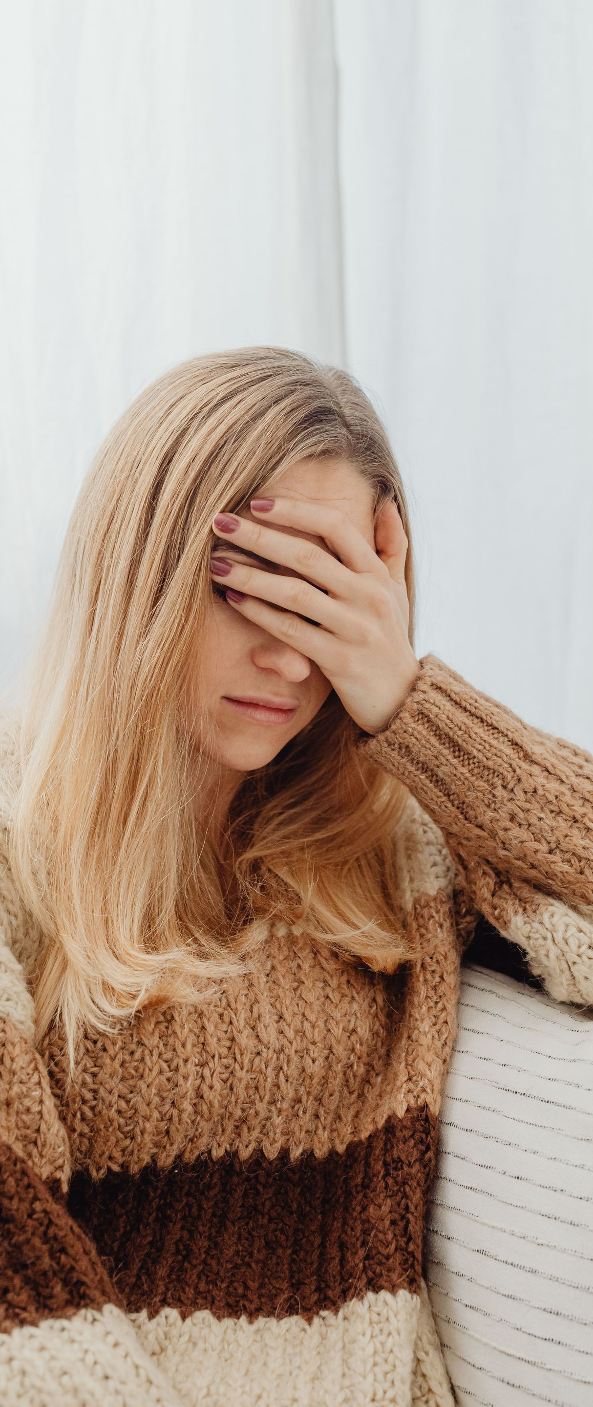 Woman in a brown sweater covering her face with her hand, sitting by a light curtain.