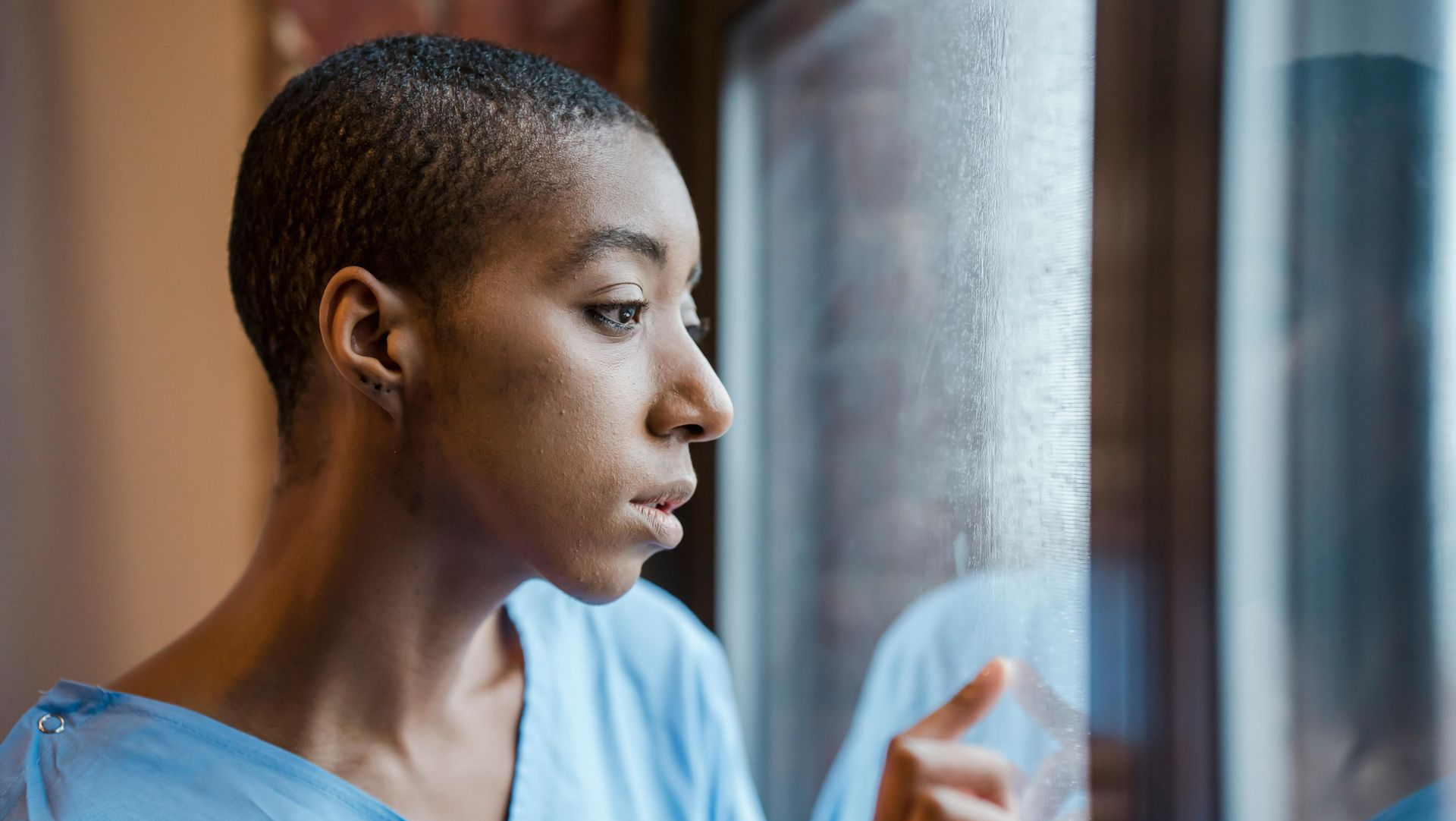 Woman in a blue scrub top looking out a window, holding a hand near the glass.