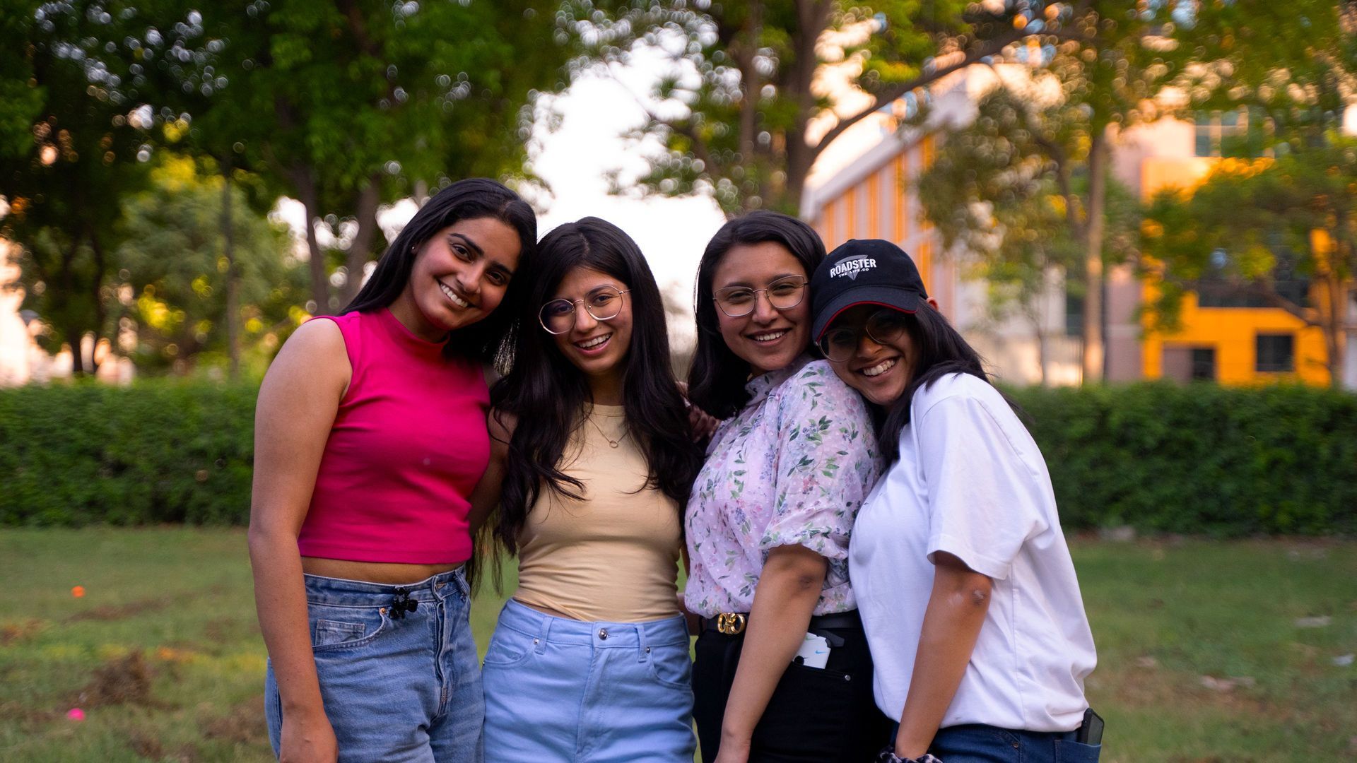 Four friends smiling and standing together outdoors in a park at sunset.