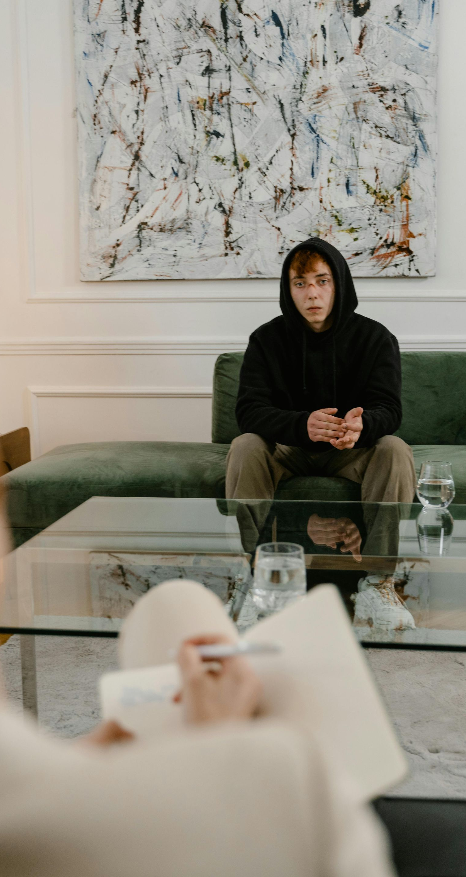 Person in a black hoodie sits on a green couch in a bright living room, with a glass coffee table in front.