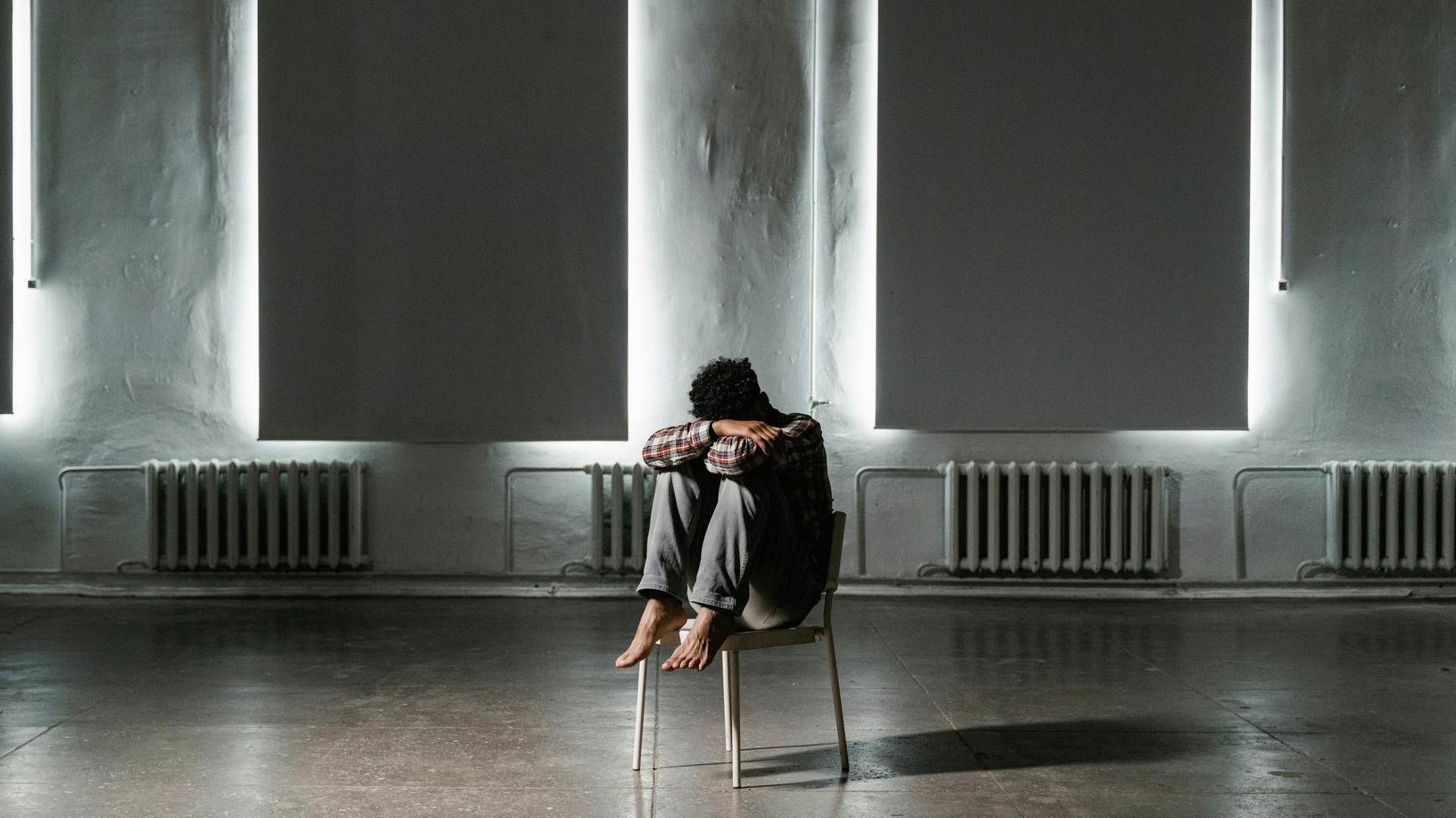 Person sitting alone on a reflective floor in a minimalist gray room with tall windows