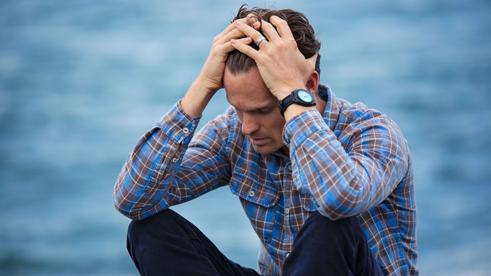 Man sitting by water with head in hands, looking distressed in a blue plaid shirt.