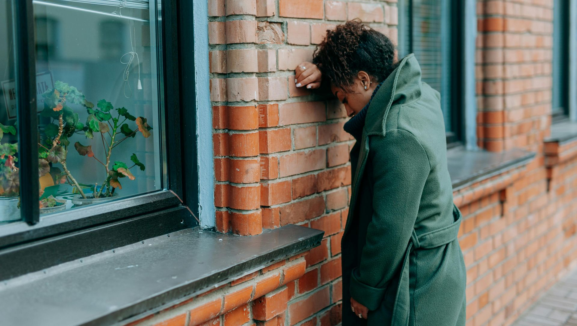 Person in a green coat leans against a brick wall beside a window, head down in distress