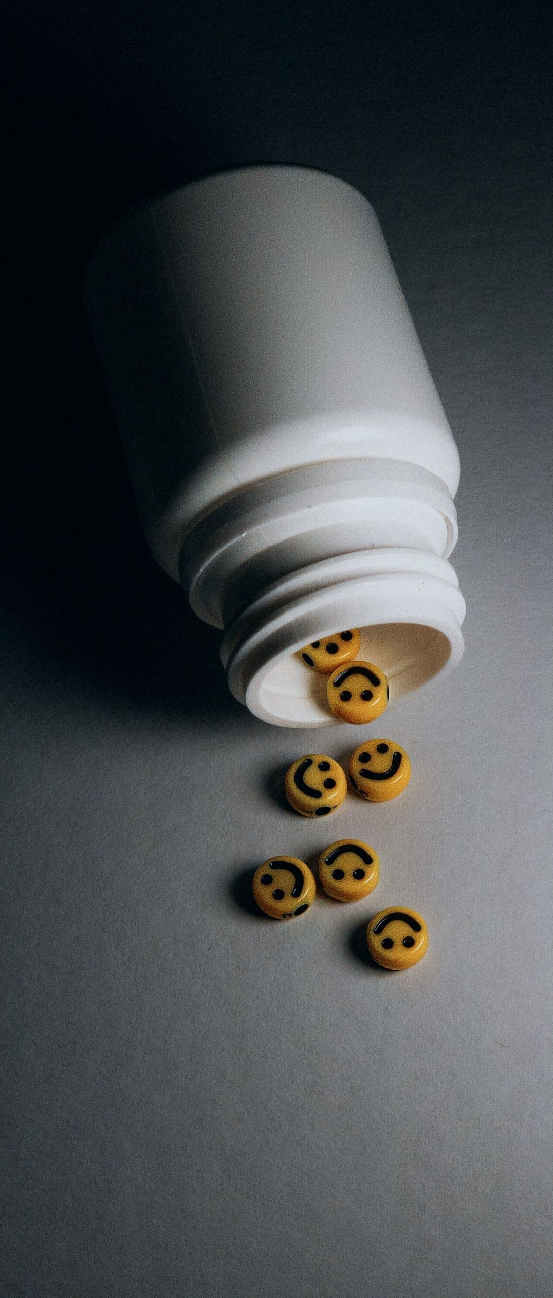 A white pill bottle tipped over, spilling smiling yellow pills onto a gray surface.