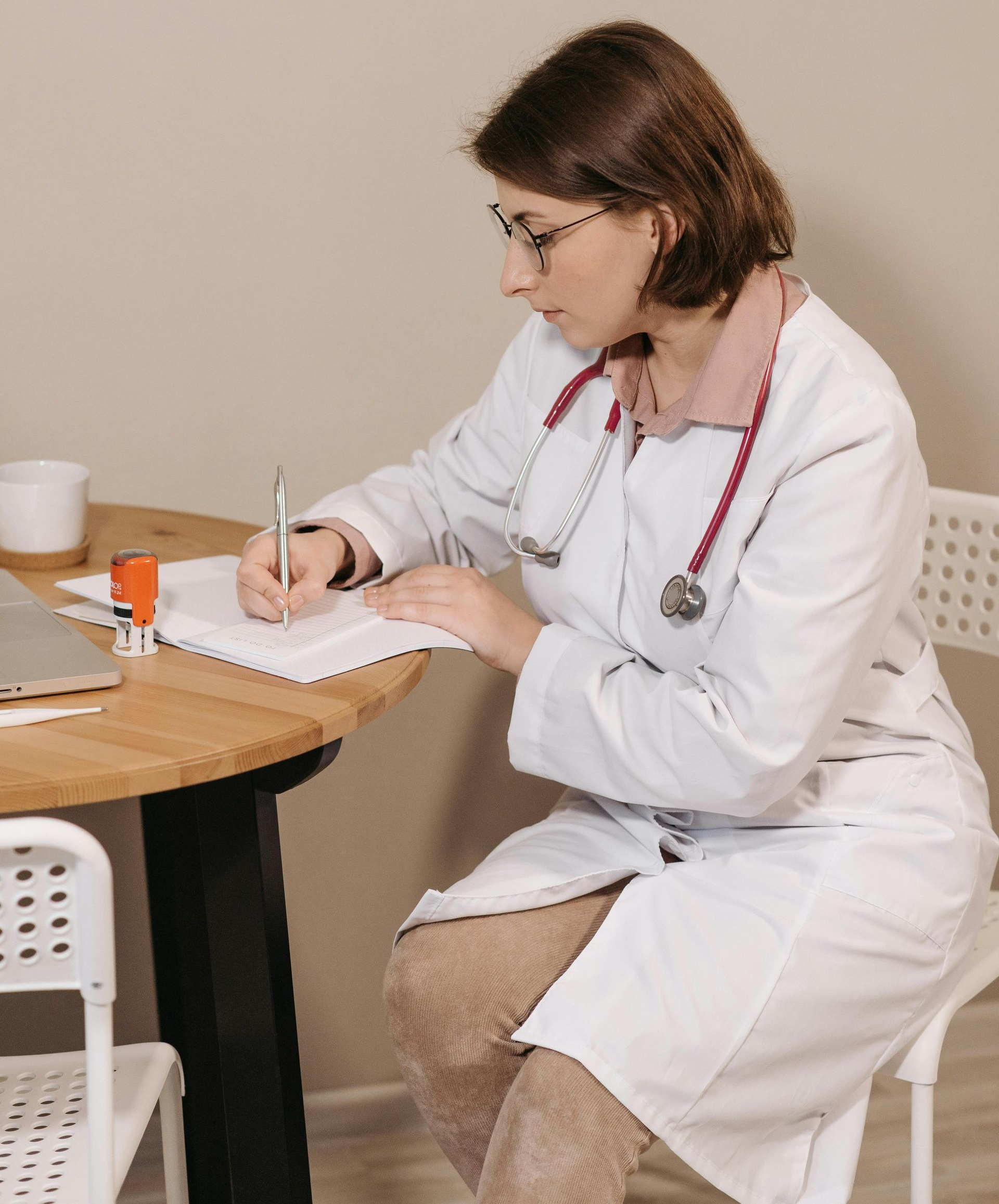 Doctor in white coat writing on a clipboard at a table in a clinic waiting area