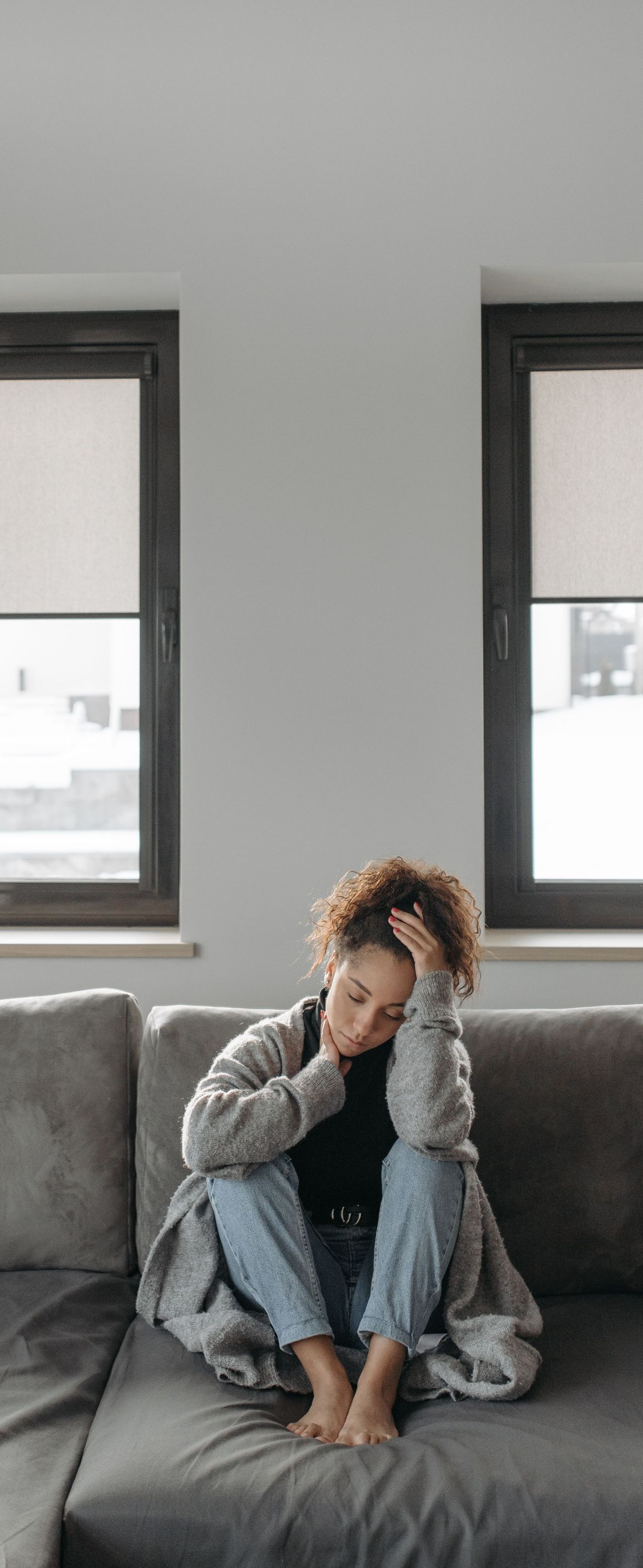 Person sitting on a gray couch, leaning forward with head in hand in a bright living room.