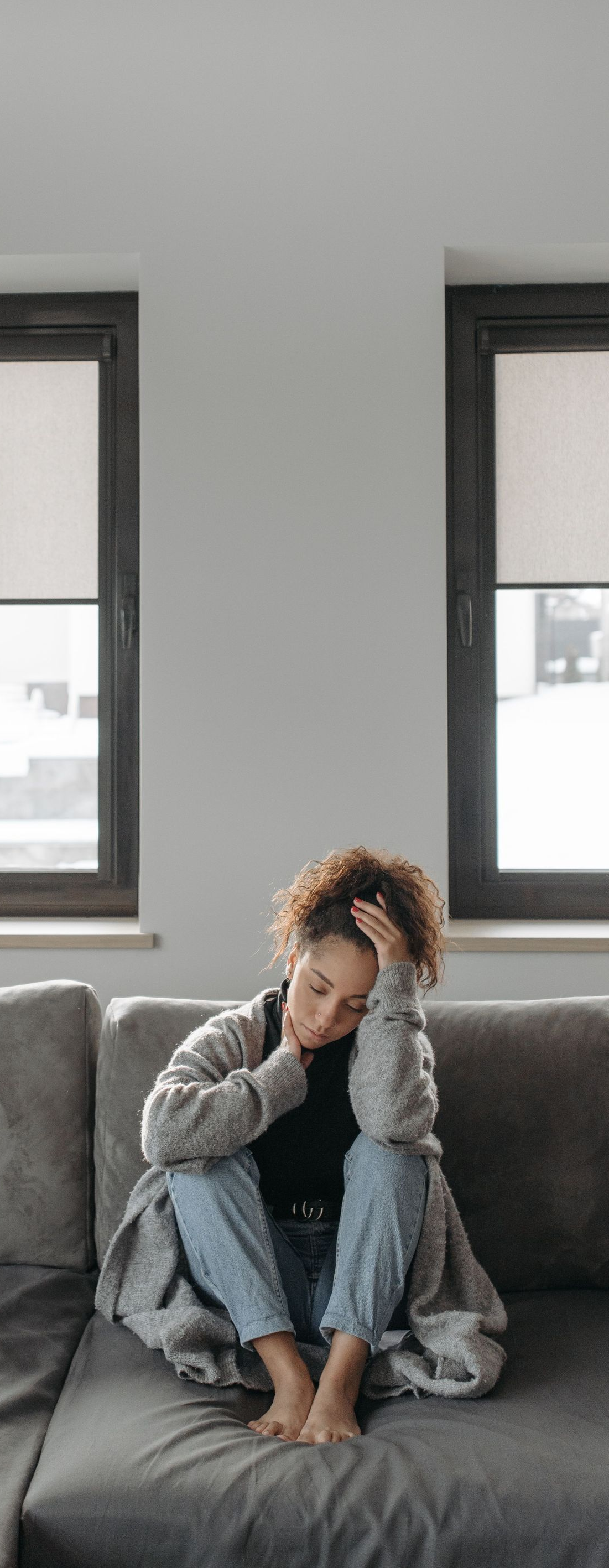 Person sitting on a gray couch, leaning forward with head in hand in a bright living room.
