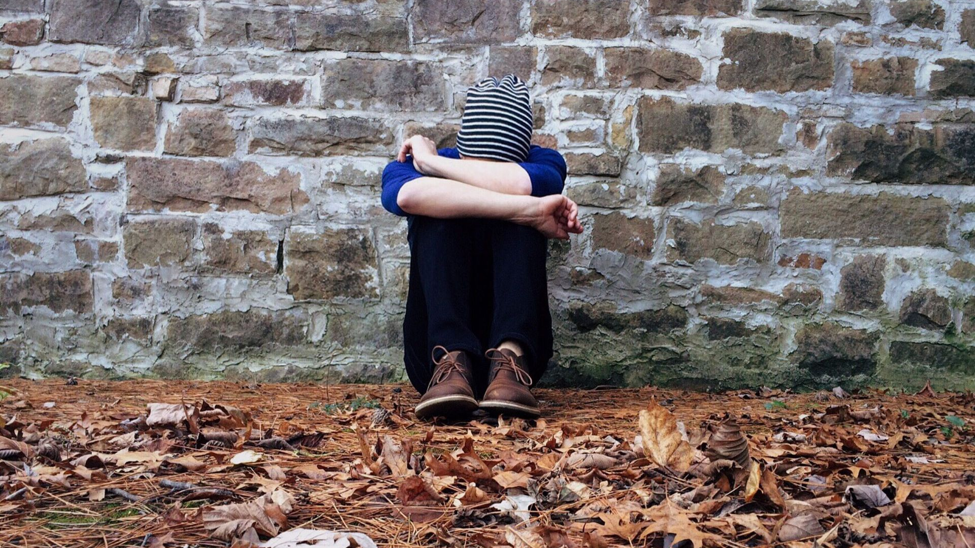 Person sitting curled up against a stone wall on a leaf-covered ground, wearing a striped hat and blue jacket.