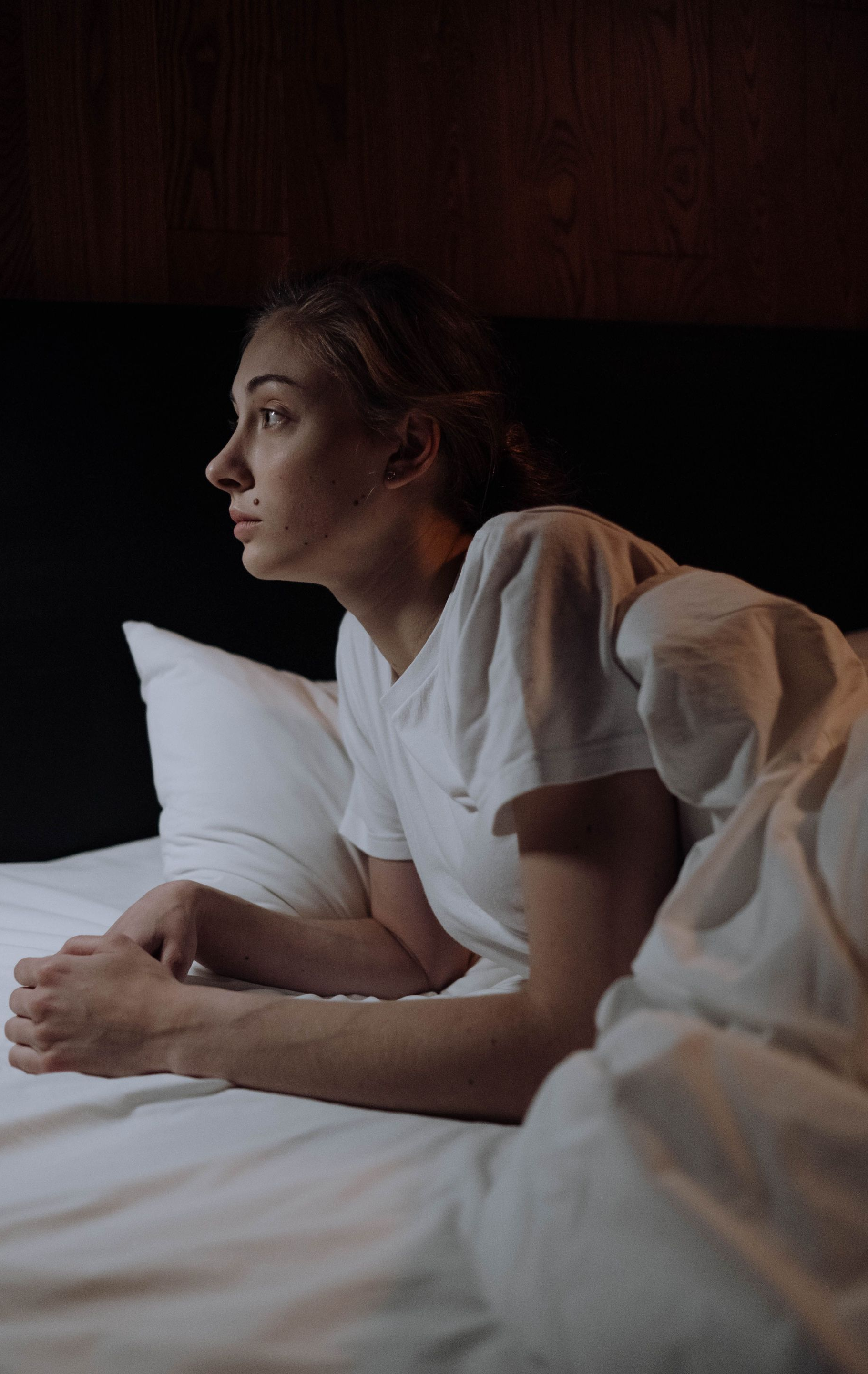 Woman in white shirt lying on a bed, looking thoughtfully to the side in dim light.