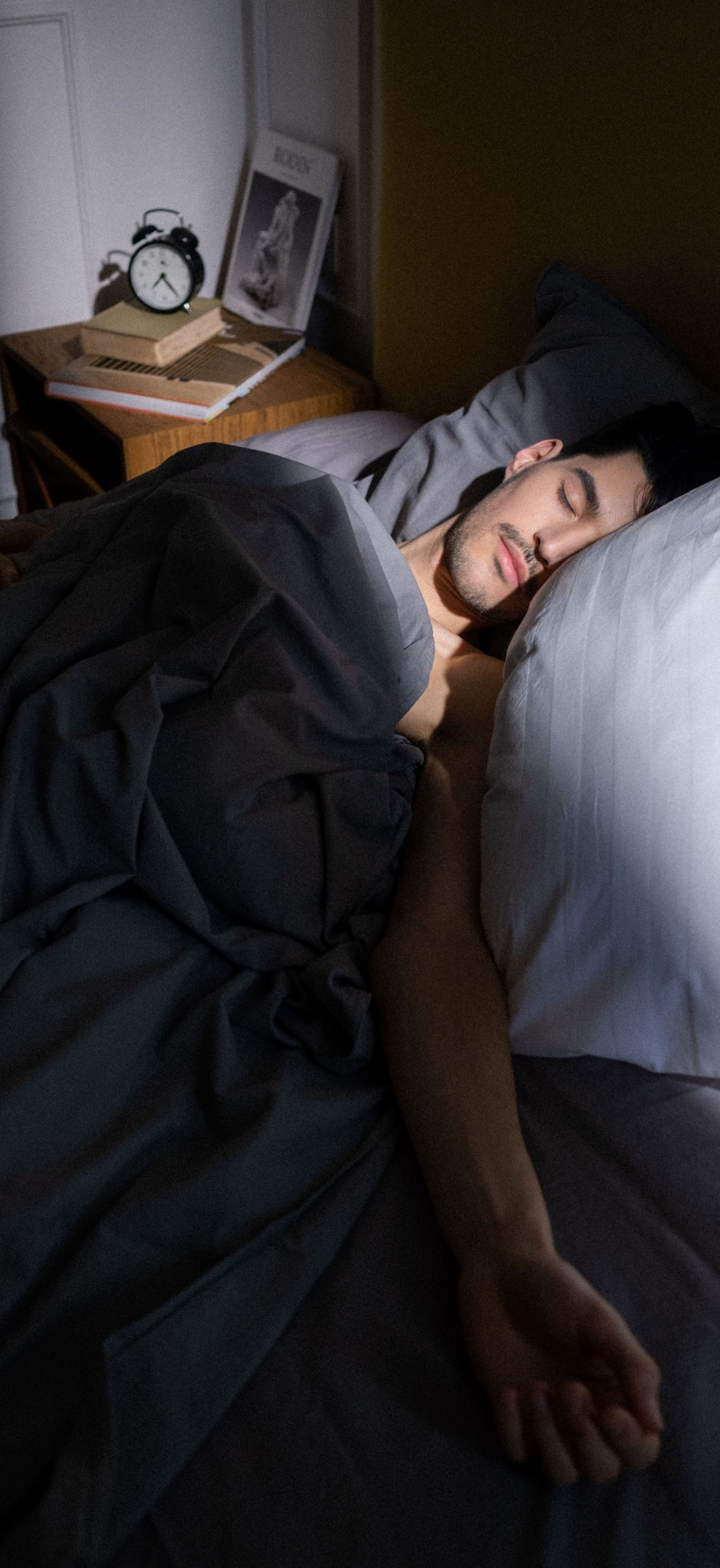 Person sleeping in bed under a dark blanket, with an alarm clock on the bedside table.