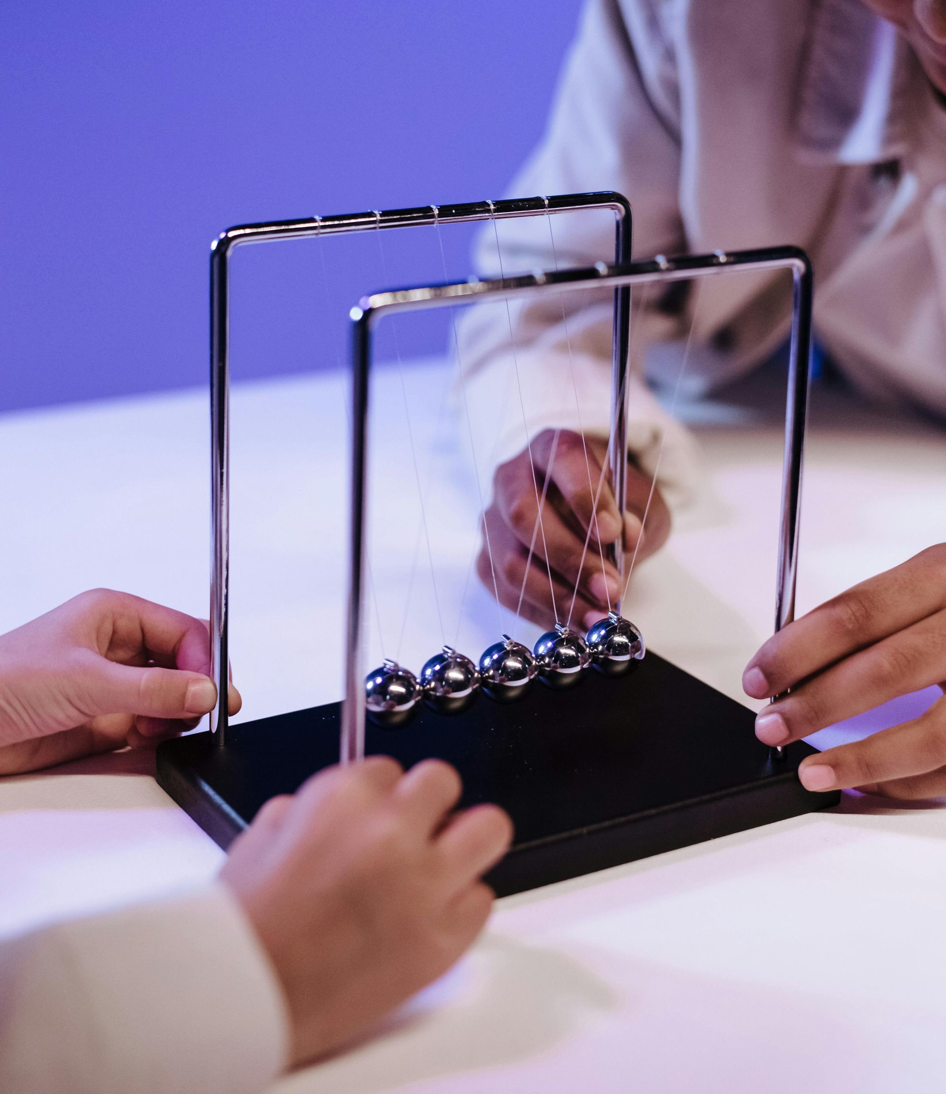 Hands playing a tabletop Newton's cradle on a table, with two people reaching in under purple lighting