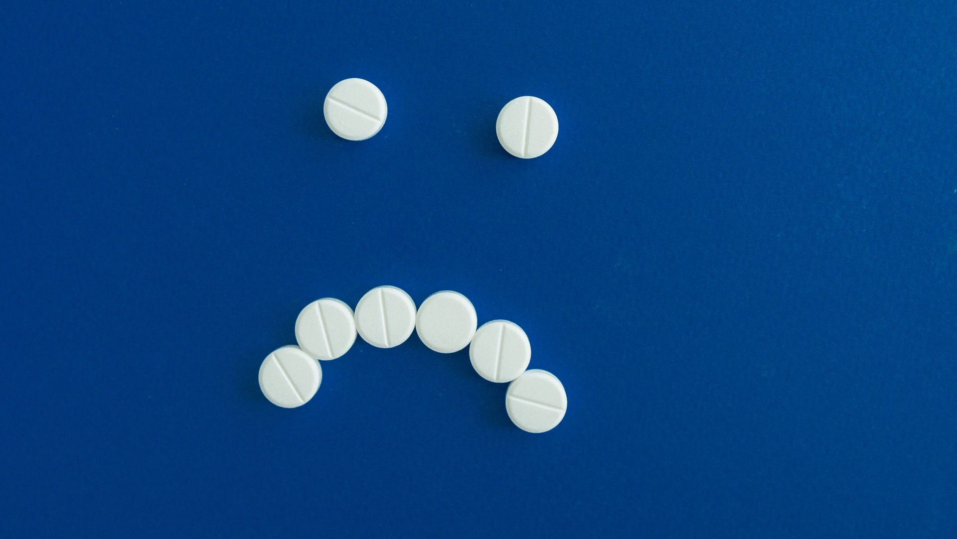 Seven white pills on a blue background arranged as a sad face.