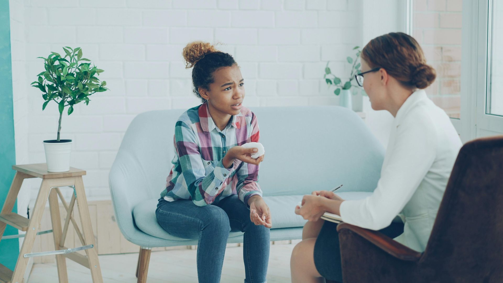 Two people talking in a bright counseling office, seated on chairs with a notebook and small plant nearby.