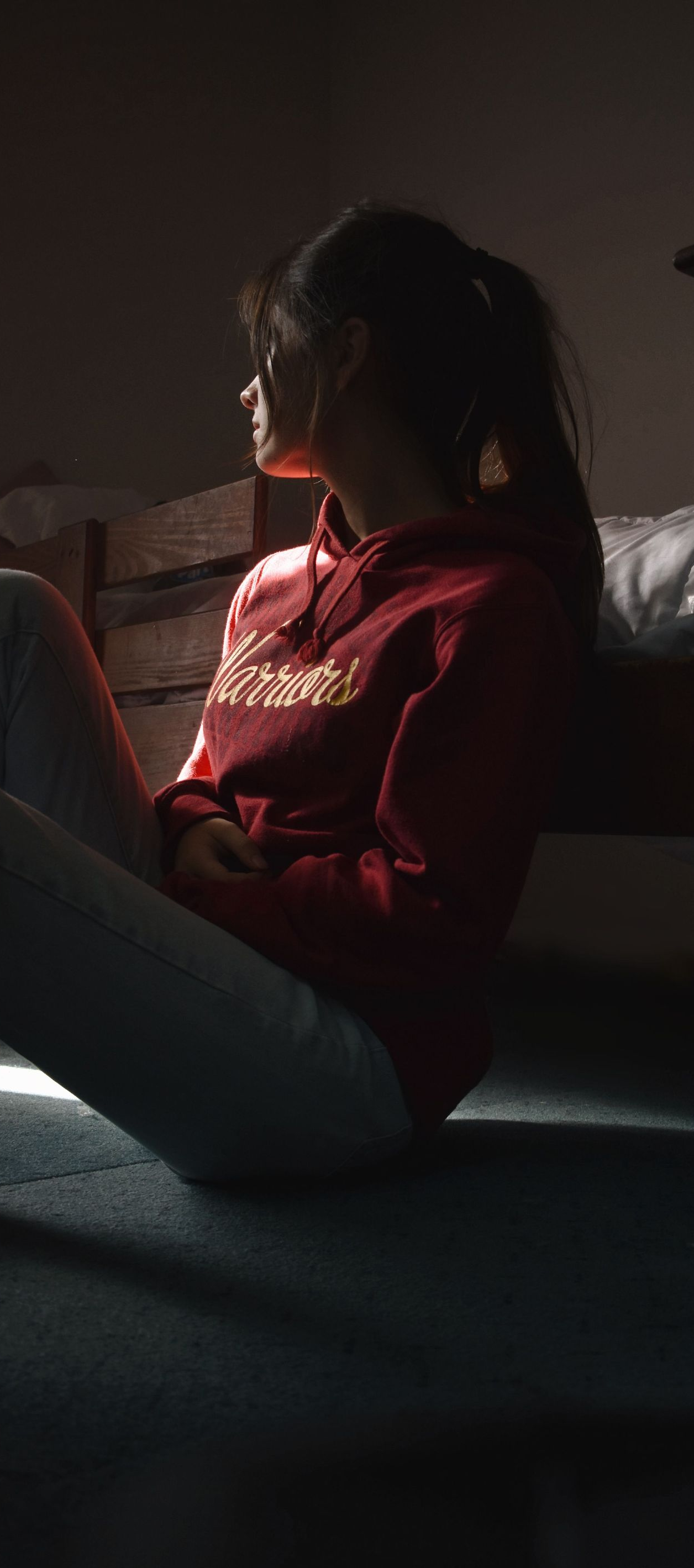 Person sitting on the floor by a staircase, lit by soft window light in a dim room.