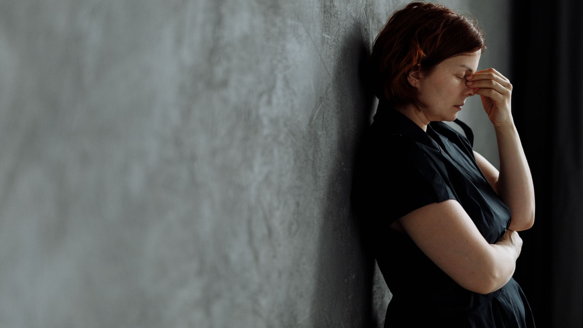 Person in a black shirt leans against a gray wall, resting a hand on their face in a contemplative pose.