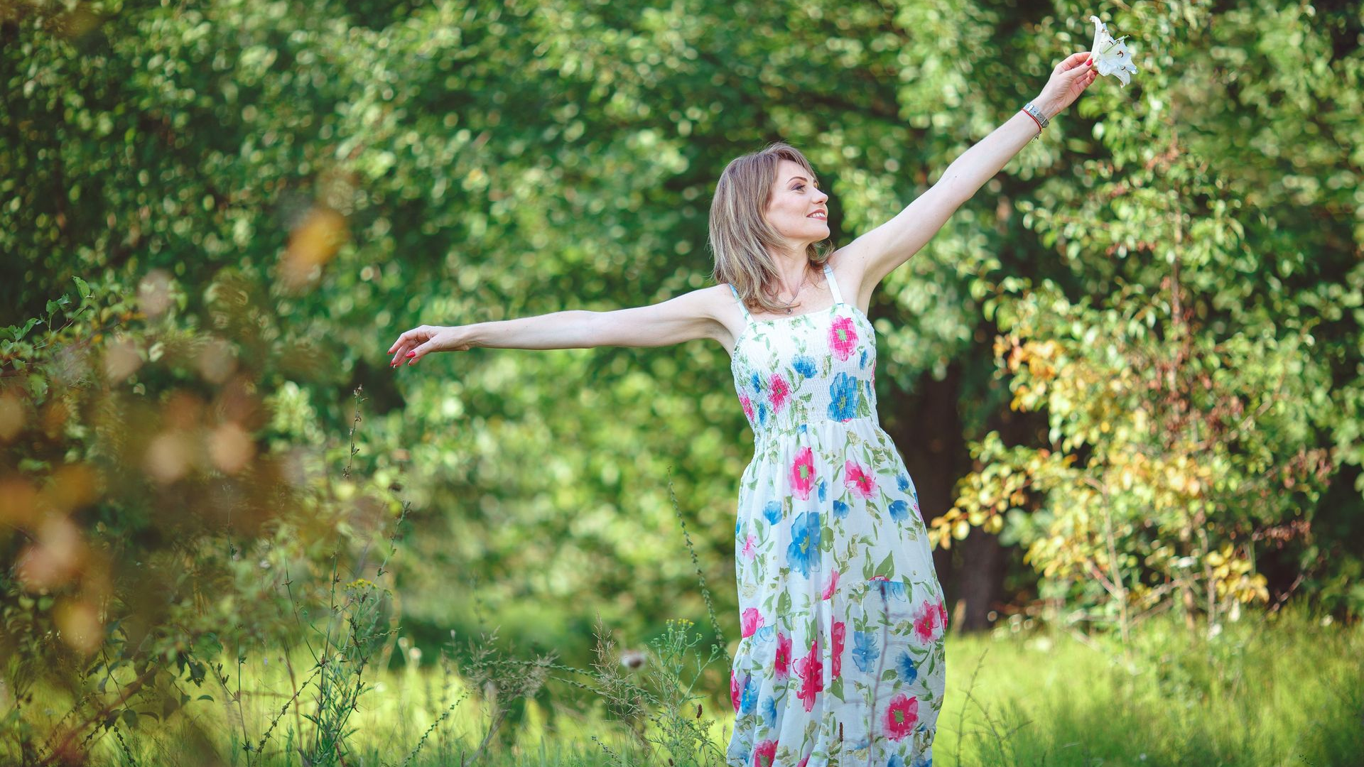 Woman in a floral dress standing in a grassy field with arms outstretched among green trees.