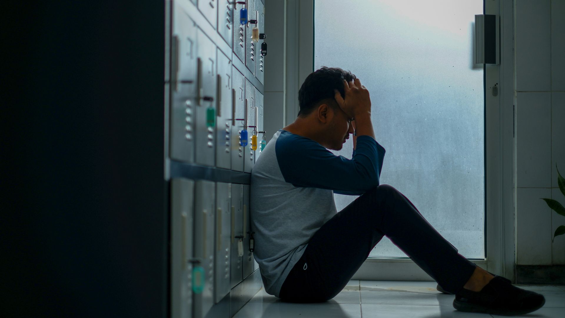 Person sitting on the floor in a hallway, head in hands, beside lockers, looking distressed
