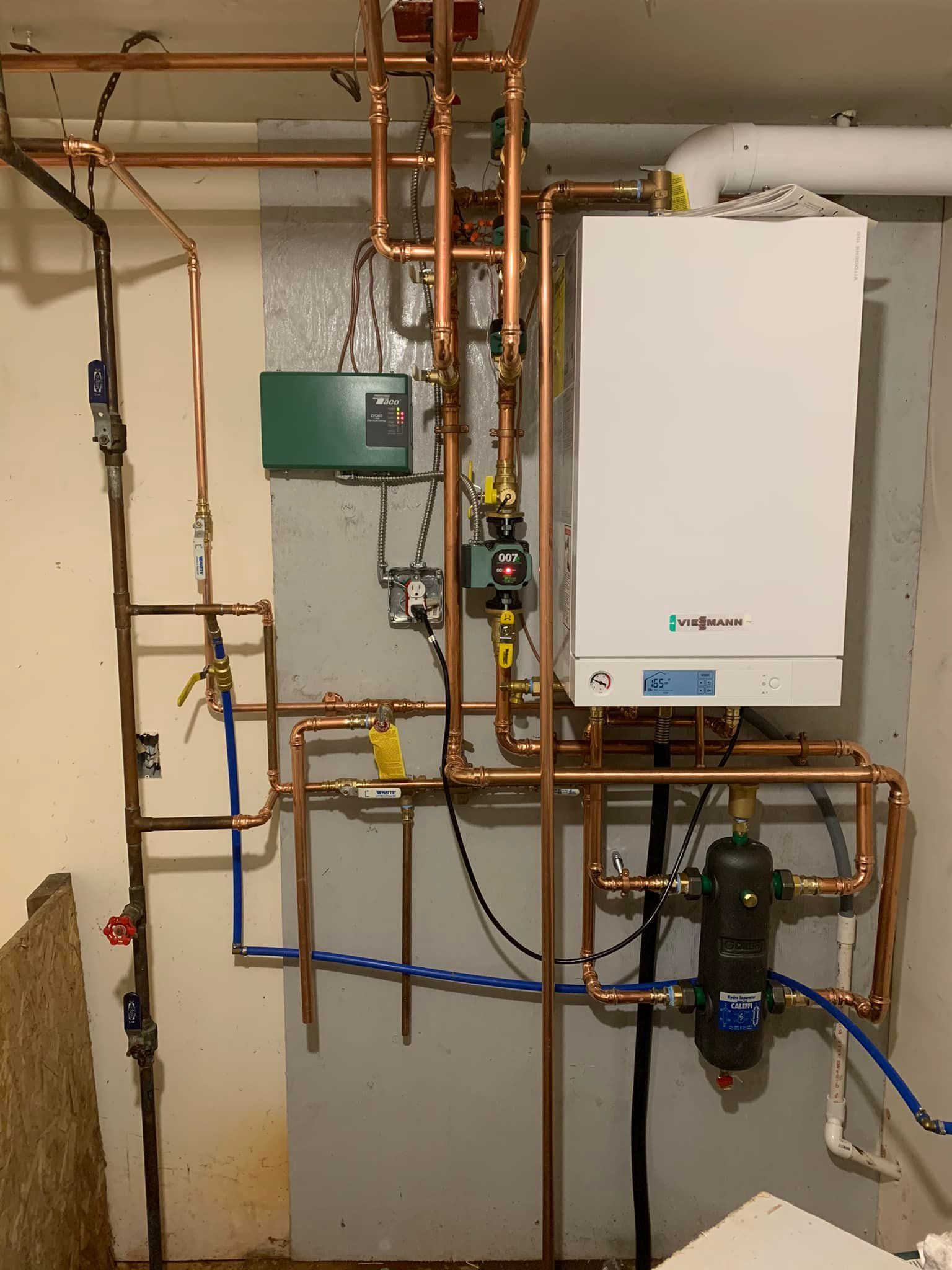 A white boiler is surrounded by copper pipes in a basement.