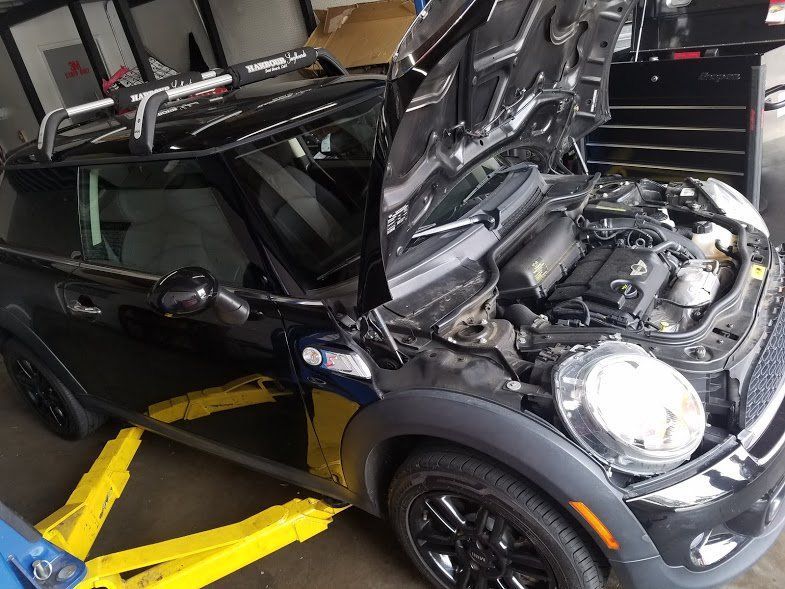 Black Mini Cooper with hood open in a garage, front left wheel strapped on a yellow lift.