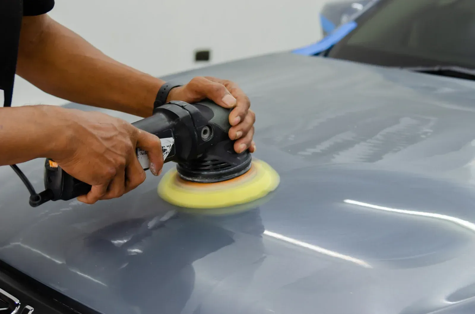Hands polishing a gray car hood with a buffer machine