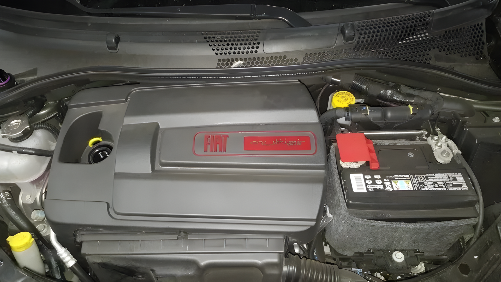 Car engine bay with black cover, red accents, battery on the right, and yellow fluid caps