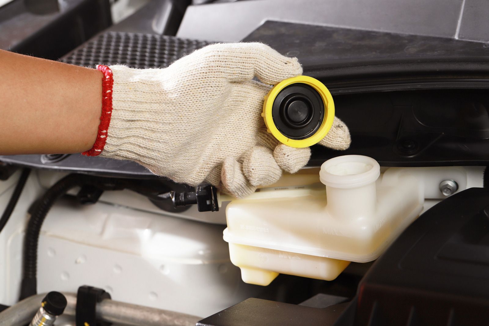 Gloved hand holding a brake fluid cap beside a car’s white fluid reservoir under the hood