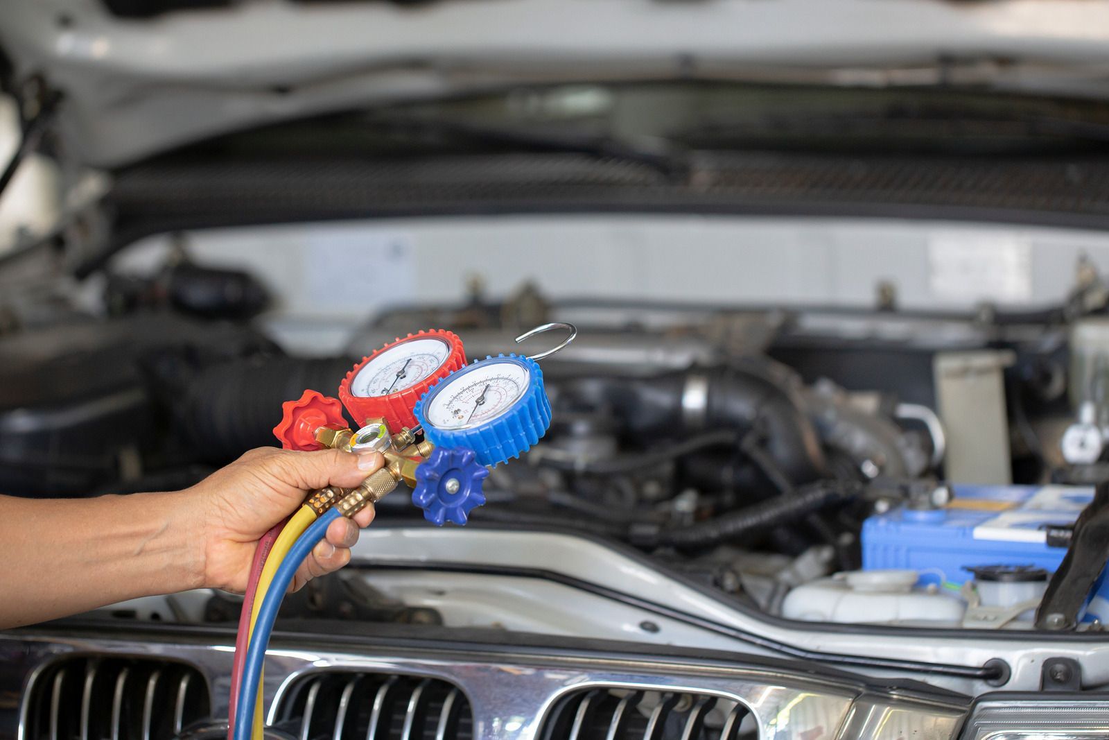 Hand holding A/C manifold gauges near car engine in a garage