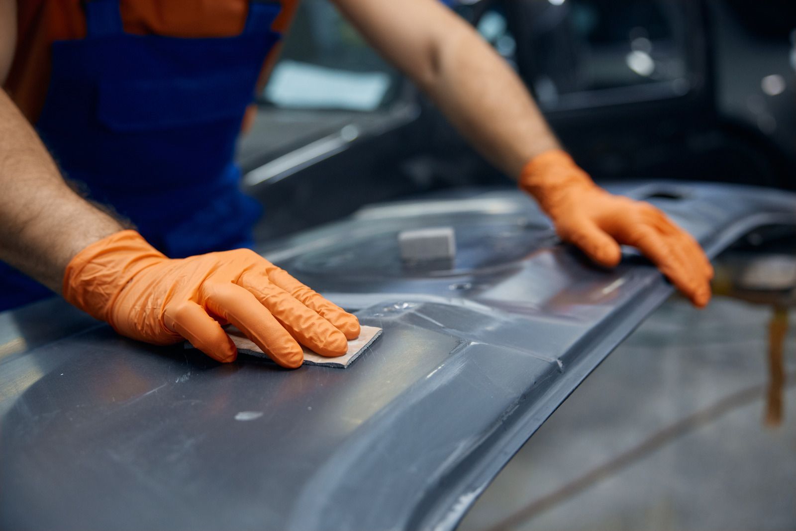 Orange-gloved hands sanding a gray car body panel in an auto repair shop