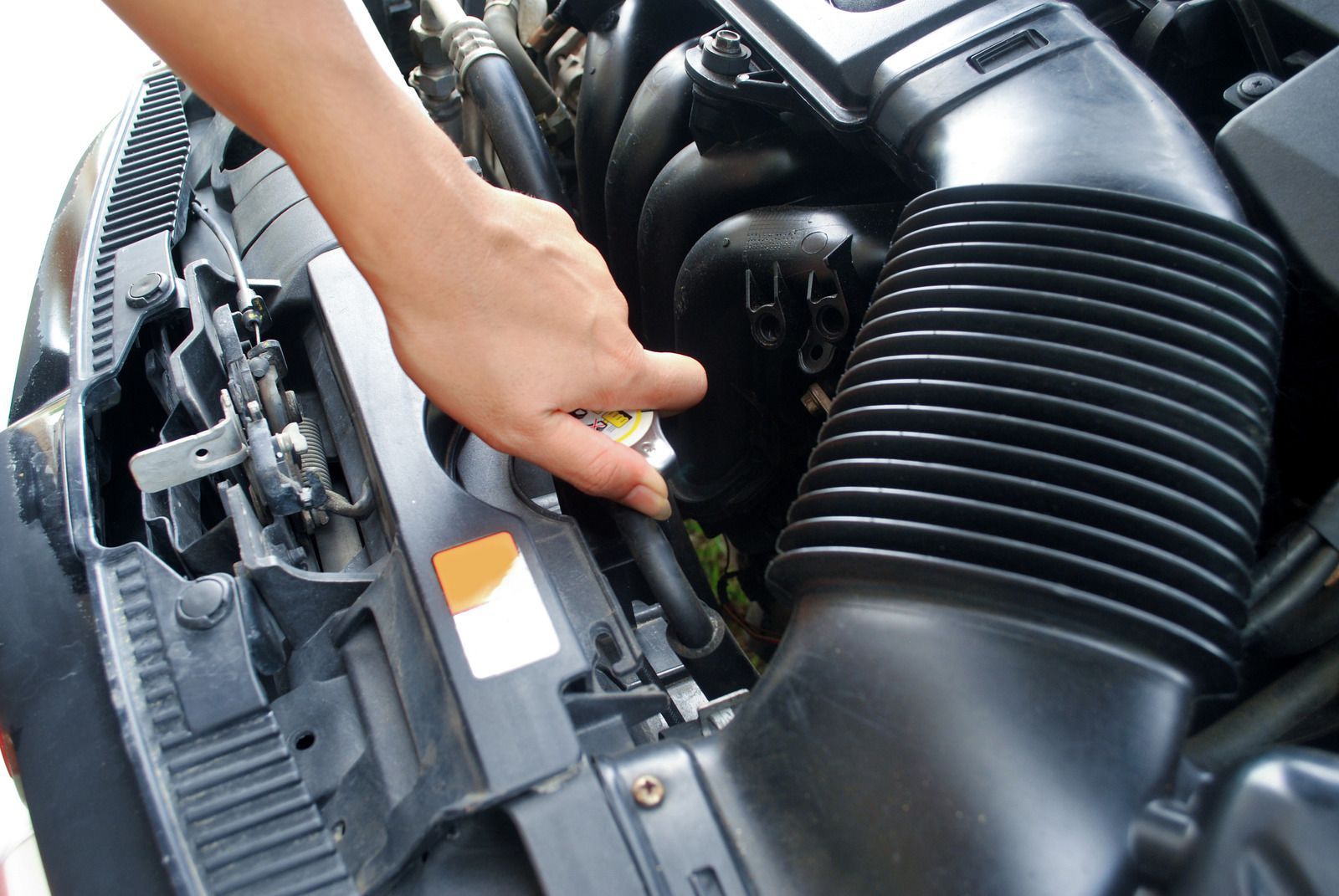 Hand checking a car engine compartment with black air intake hose and orange label