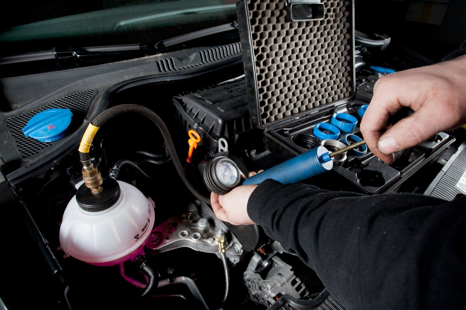 Hands working under a car hood, using tools near the engine and coolant reservoir.