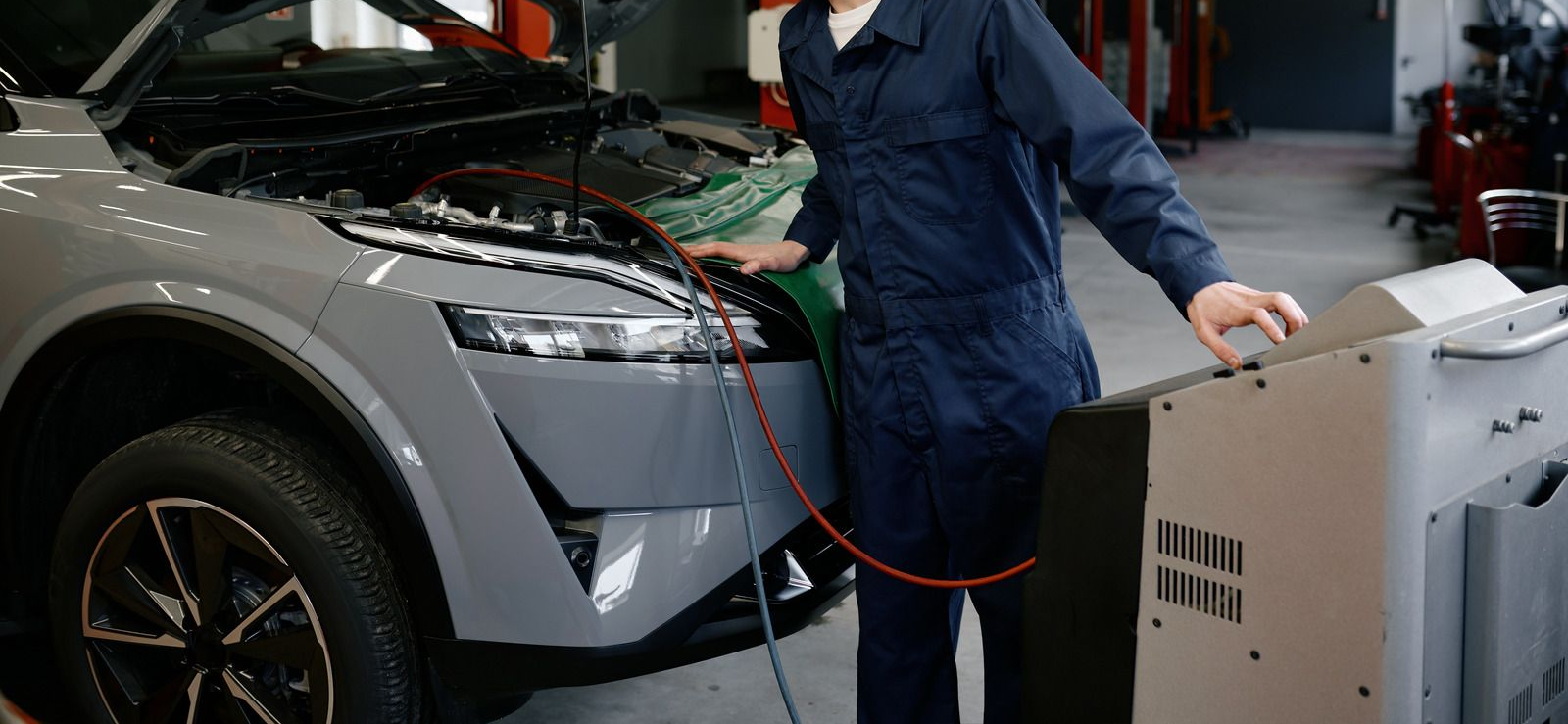 Person in blue coveralls checking the front of a gray car beside test equipment in a workshop