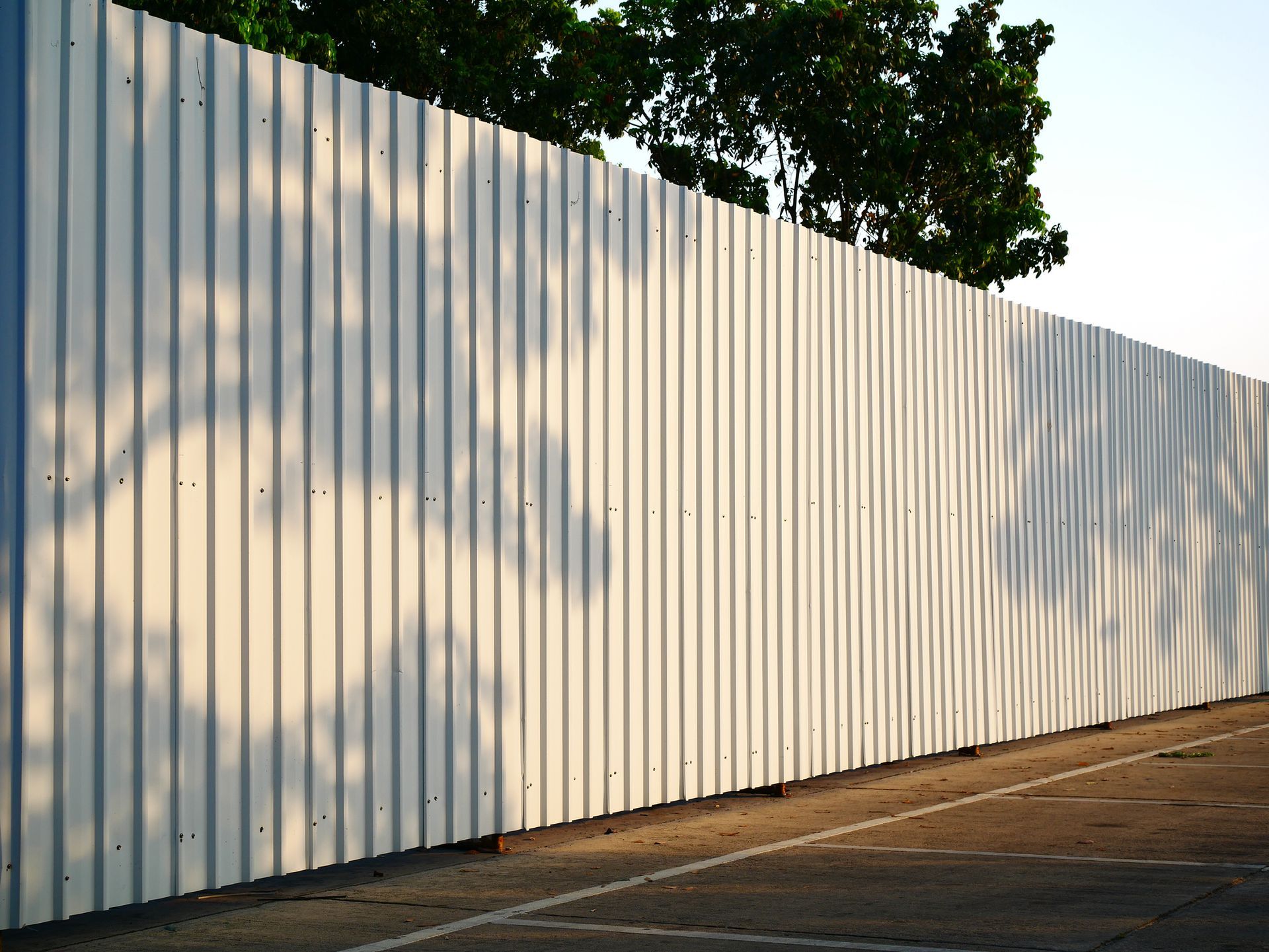 White corrugated metal construction fence along sidewalk casting tree shadows in daylight sun light.