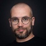 Een kale man met baard en bril draagt een zwart shirt.