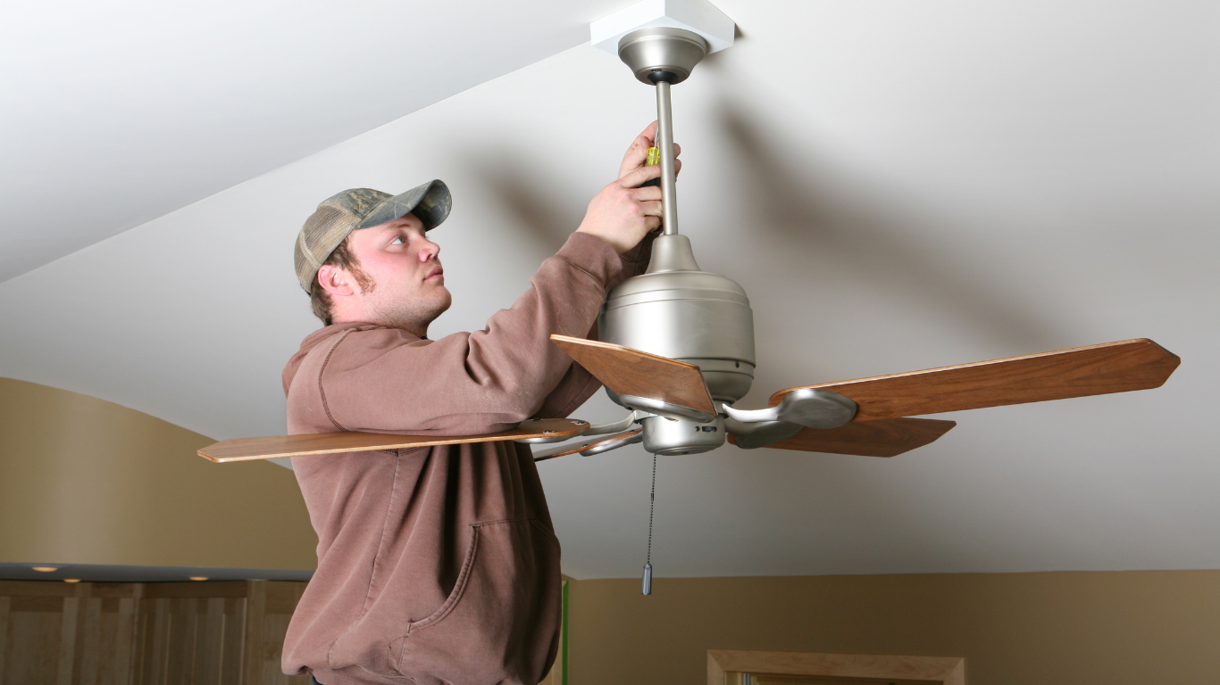 Man in a brown hoodie and cap installing a ceiling fan in a room.