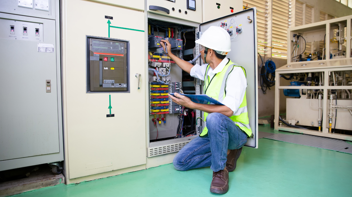 Electrician kneeling, examining wiring in open electrical panel, wearing a hard hat and safety vest in a plant.
