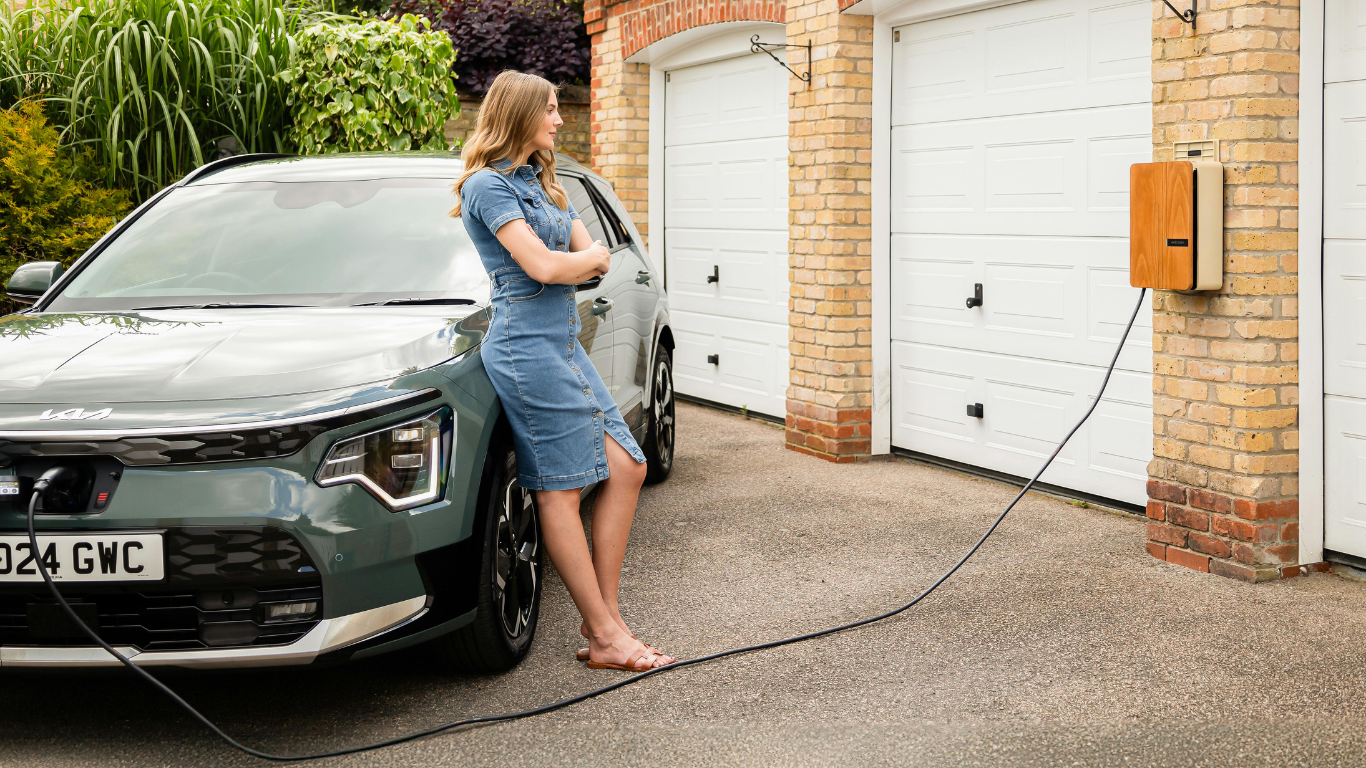 Woman leans on an electric car charging in front of a garage.