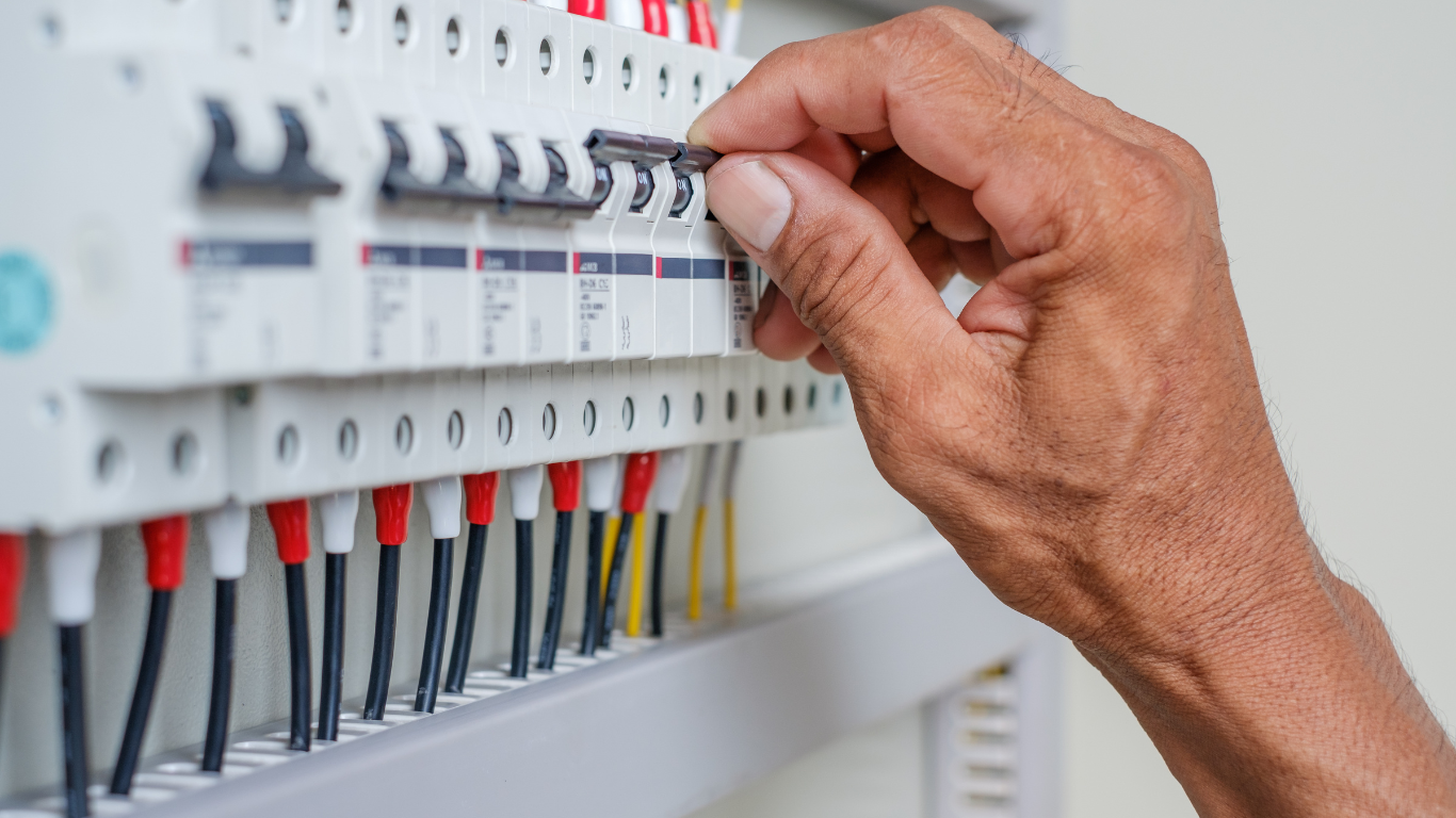 Person's hand flipping a circuit breaker switch in a panel with many switches and colored wires.