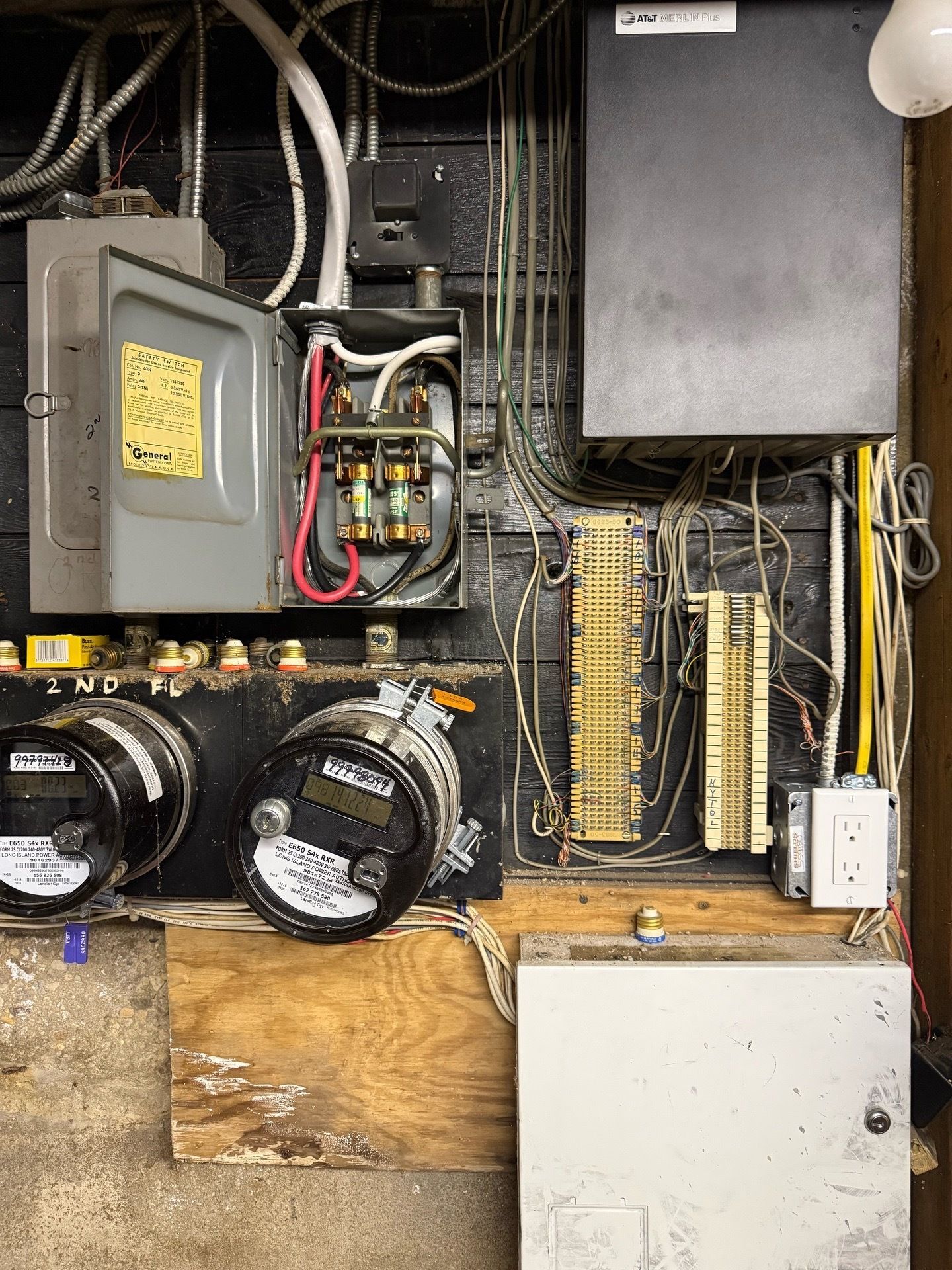 Two electricians in blue shirts and caps working on a circuit breaker panel. One points, the other tests wires.