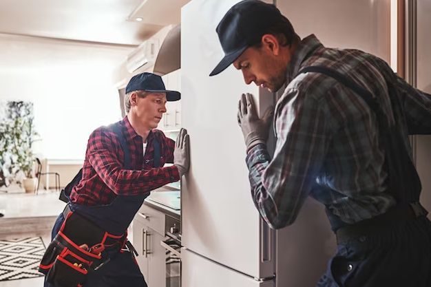 Two men in work clothes pushing a refrigerator indoors.