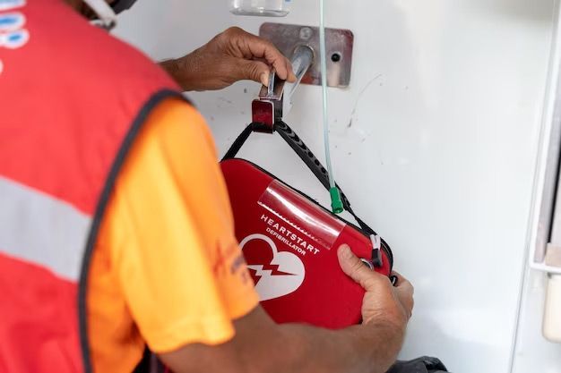 Person in orange vest removing AED from a wall mount. Red case with heart symbol.