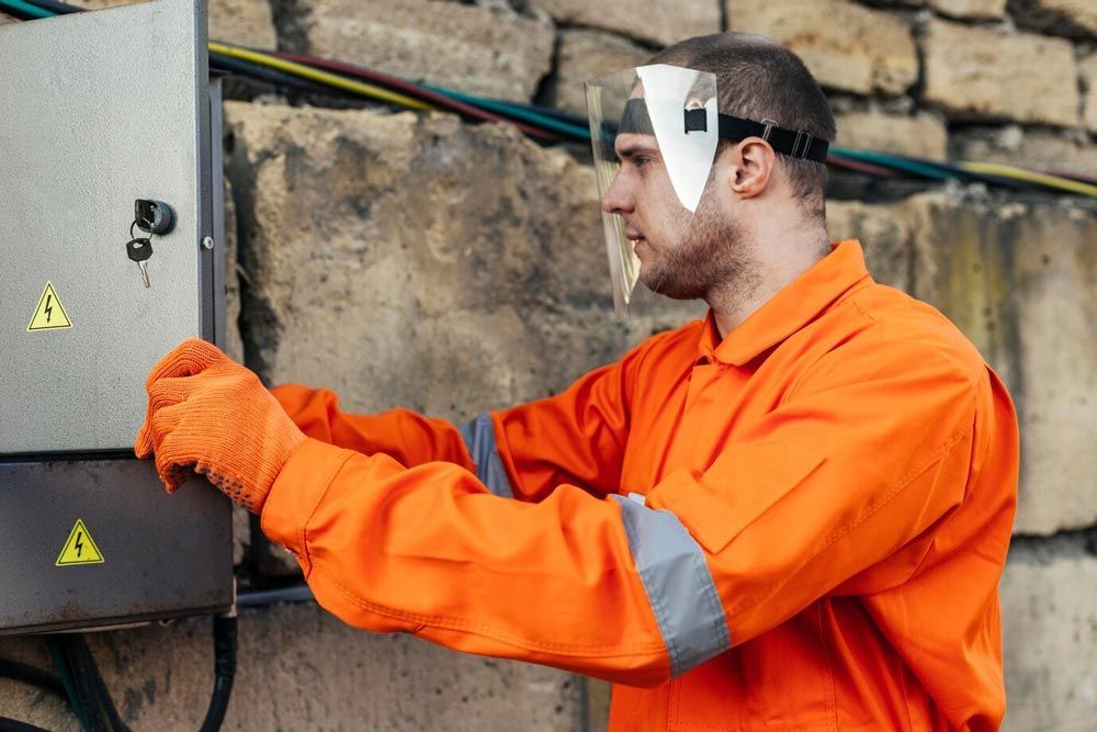 Electrician in orange workwear with face shield and gloves working on a gray electrical box outdoors.
