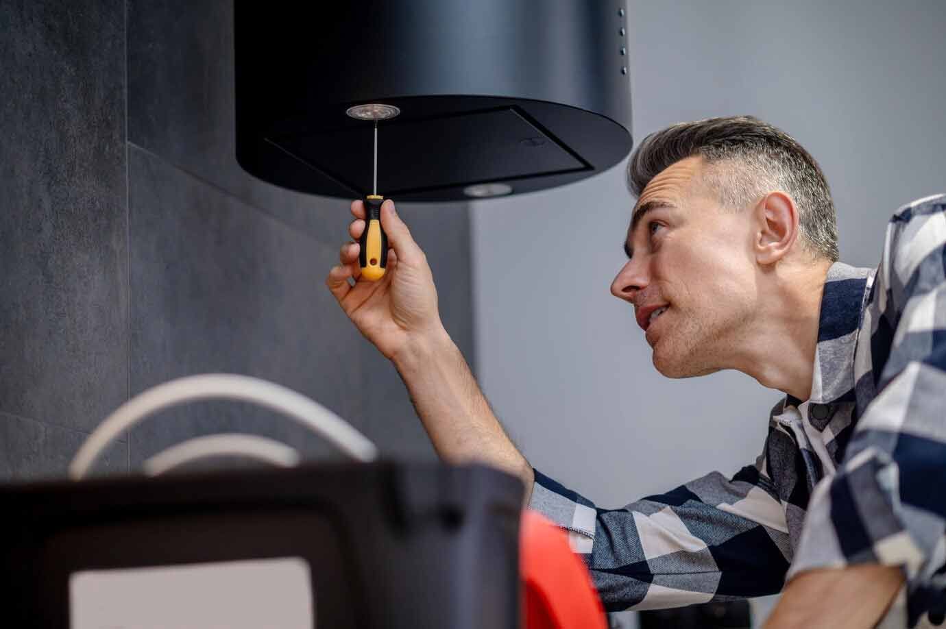 Man in checkered shirt using a screwdriver to install a black kitchen hood against a gray wall.