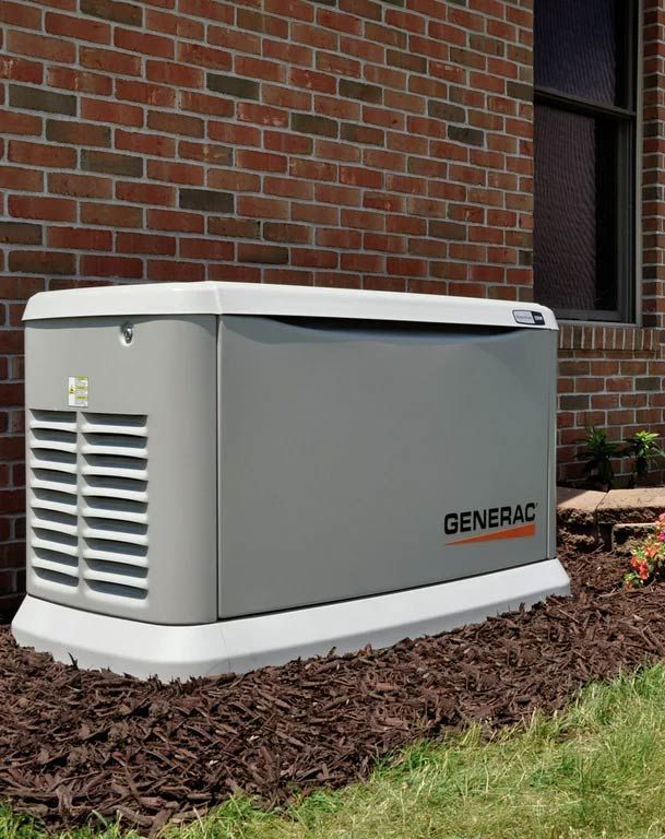 Generac home generator, gray with white base, by a brick wall and window, sitting in mulch.