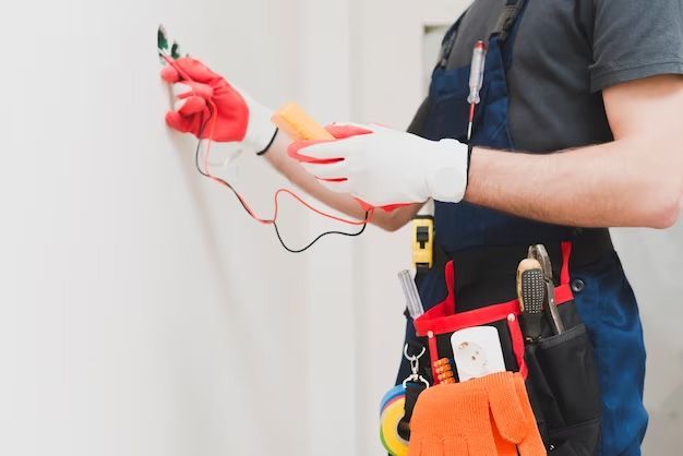 Electrician in blue overalls and gloves tests a wall outlet with a multimeter.