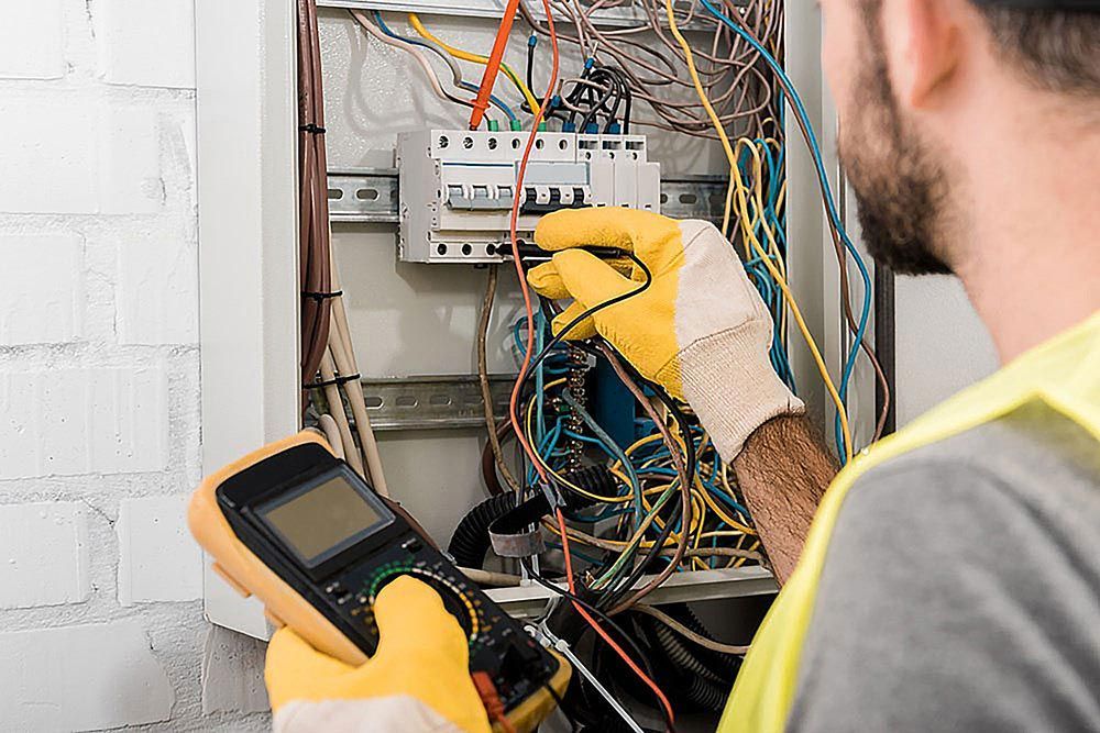 Electrician using multimeter inside an electrical panel, wearing gloves and safety vest.