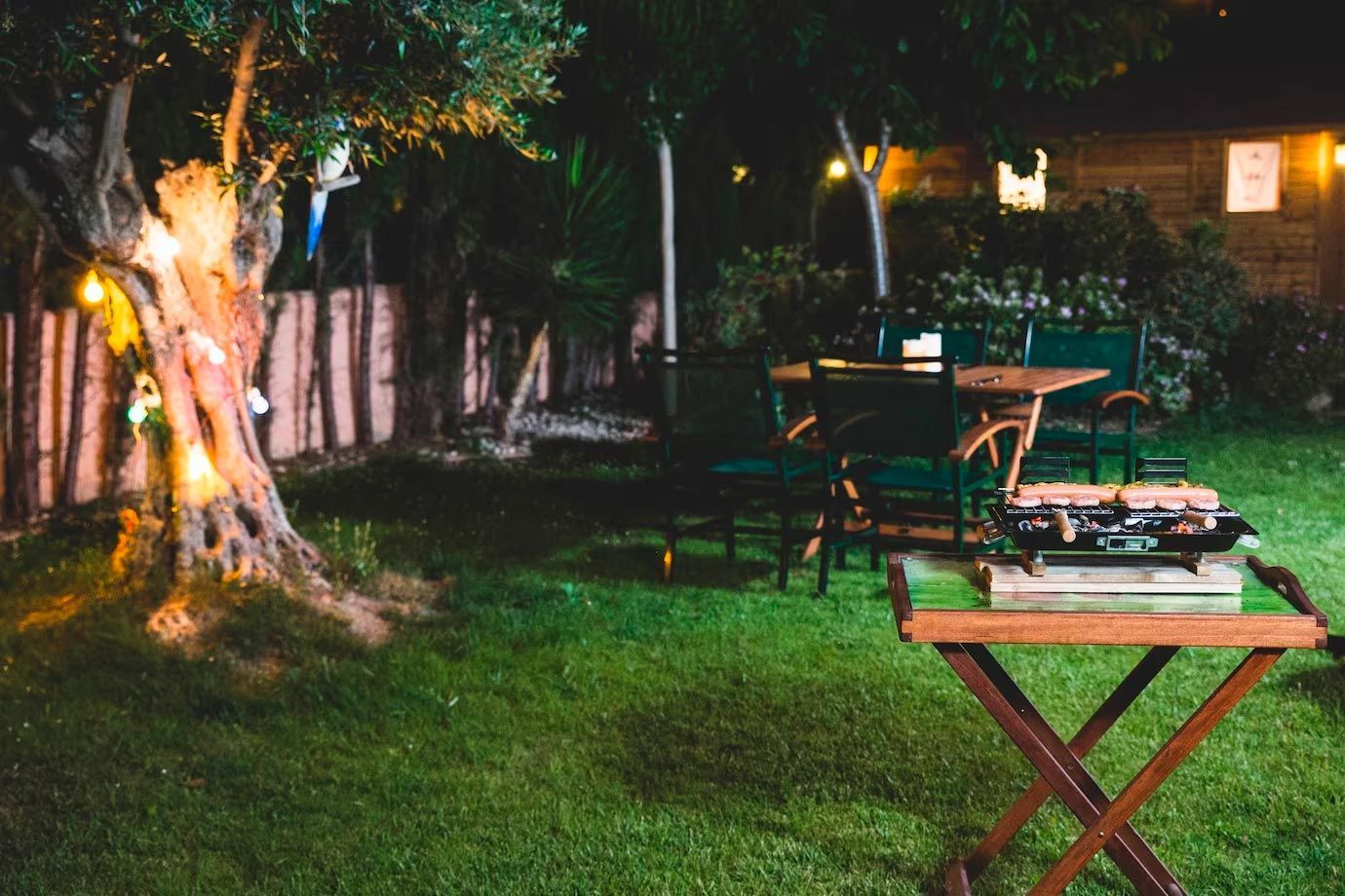 Nighttime backyard scene with a grill, table, chairs, and a lit-up tree.