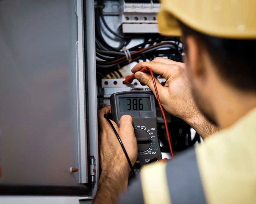 Electrician in yellow hard hat tests electrical panel with a multimeter, displaying